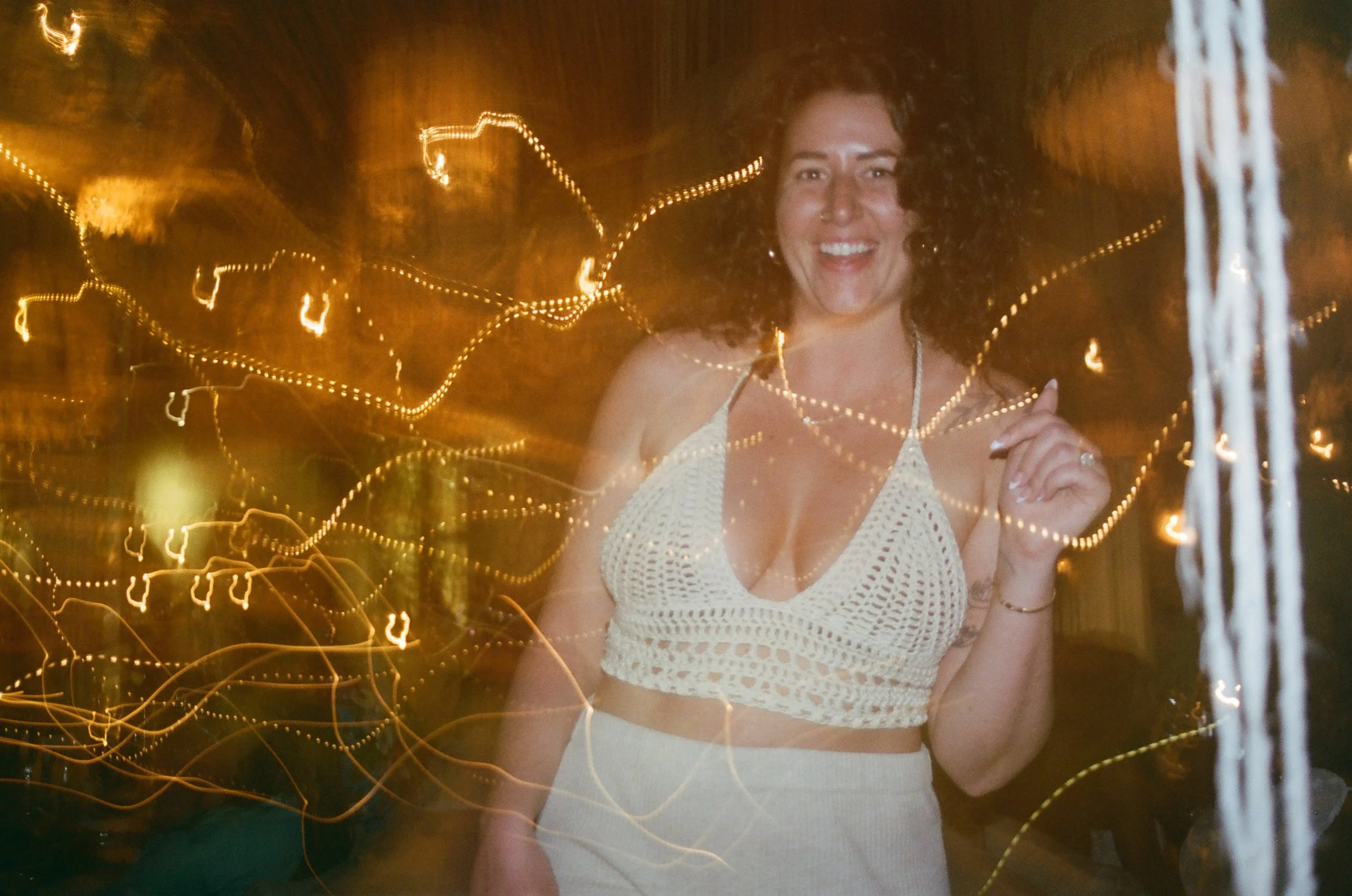 A woman in a white crochet top smiling at night with string lights creating a bokeh effect around her.