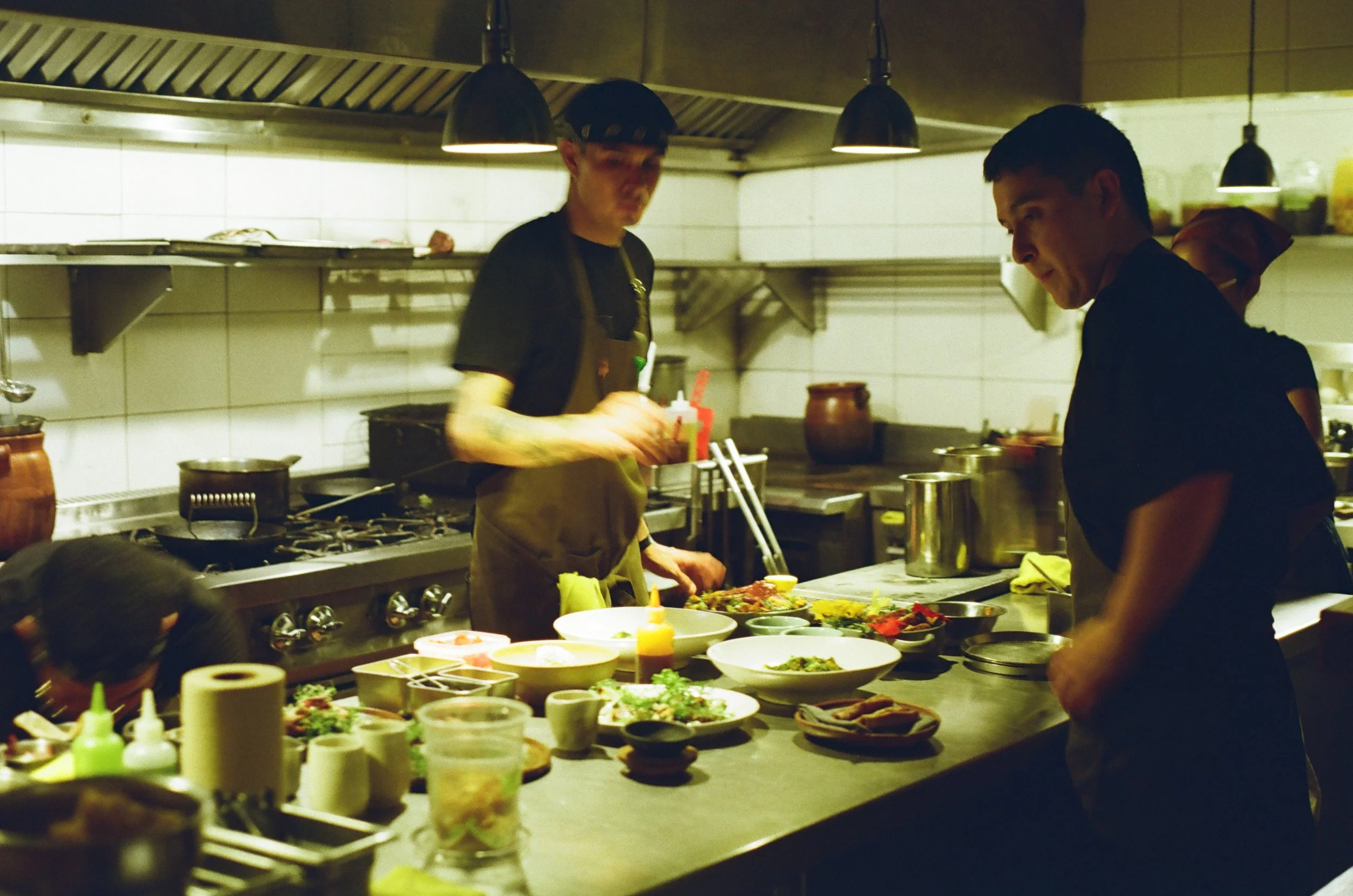 Two chefs working in a professional kitchen, preparing and arranging dishes on a counter filled with various bowls of ingredients and garnishes.