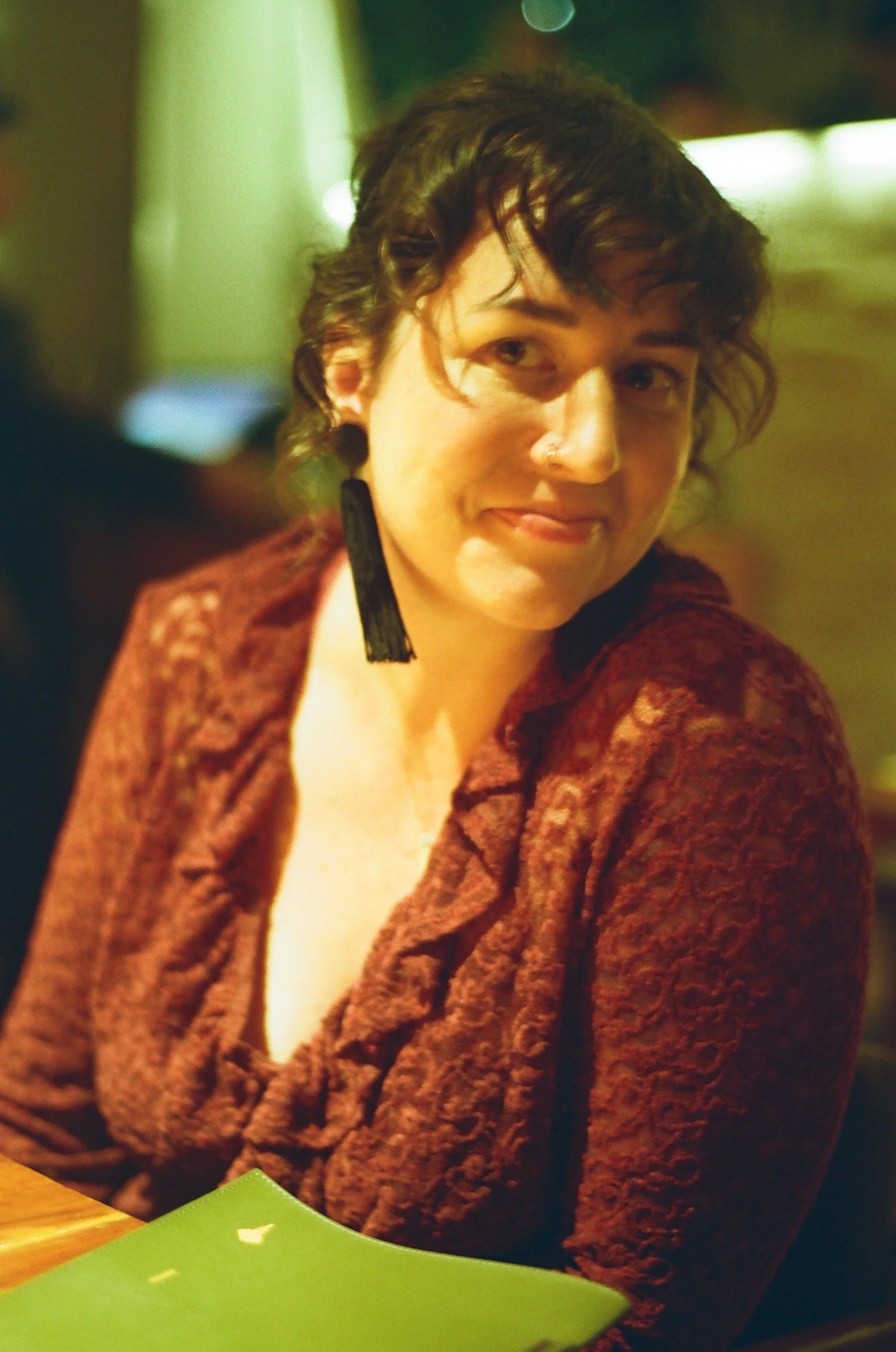 A person with short curly hair and a nose piercing wearing a patterned red jacket, looking to the side with a slight smile.