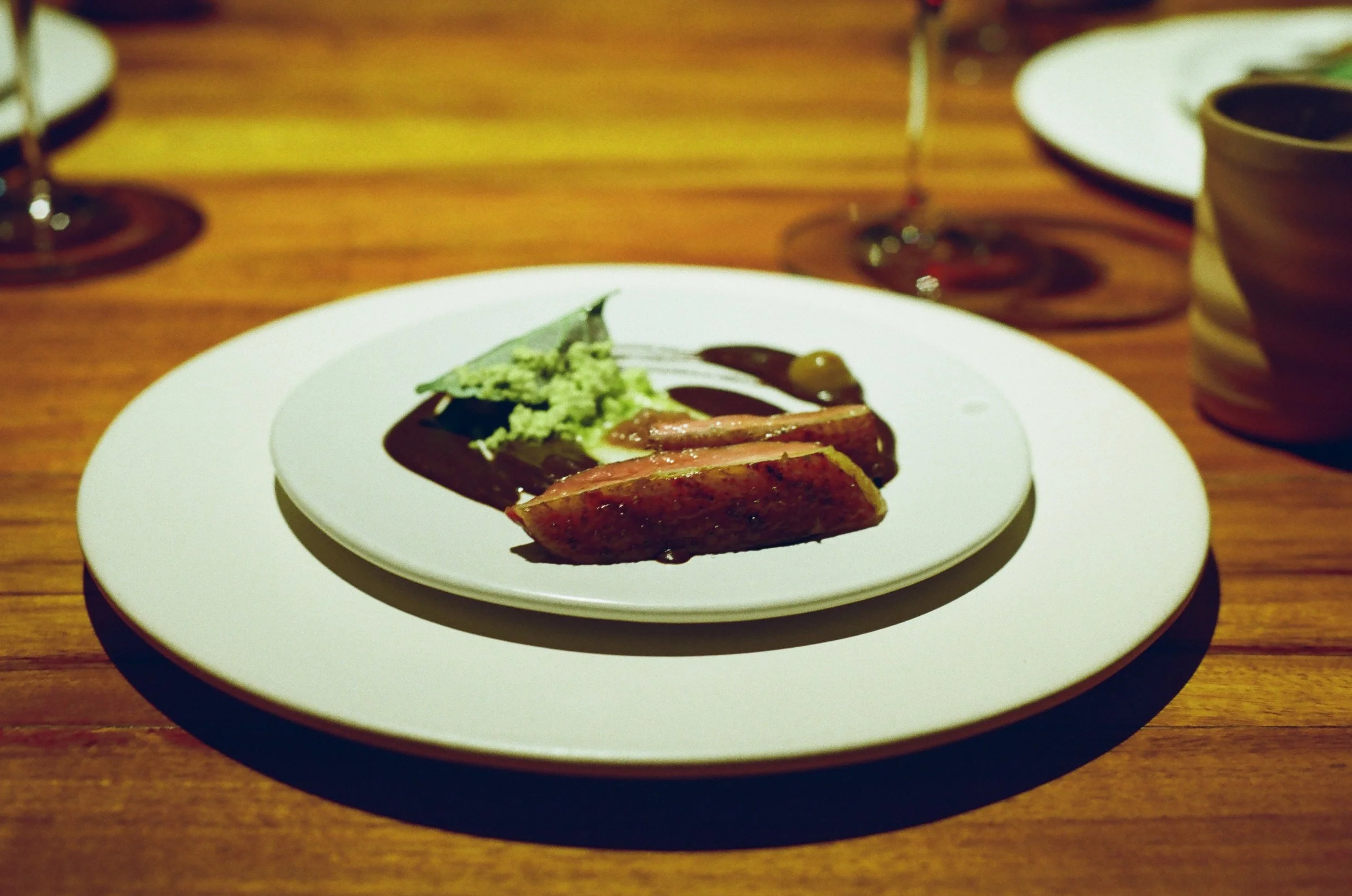 A white plate with a piece of meat topped with green guacamole and dark sauce, and slices of meat on the side, placed on a wooden table with two wine glasses and a cup in the background.