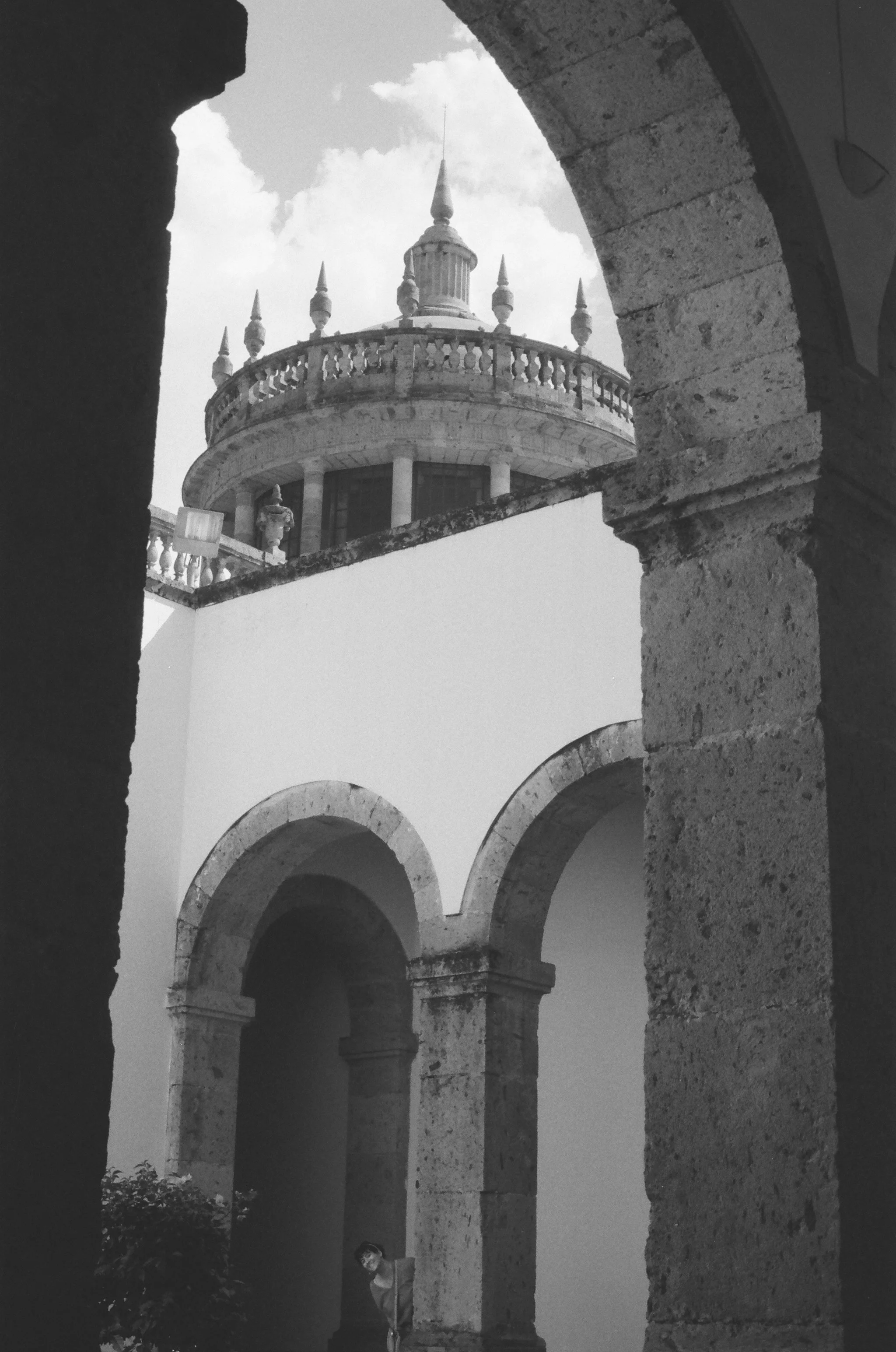 Black and white photo of a stone structure with arches and a balcony, viewed through an archway, with a person partially visible near the bottom.