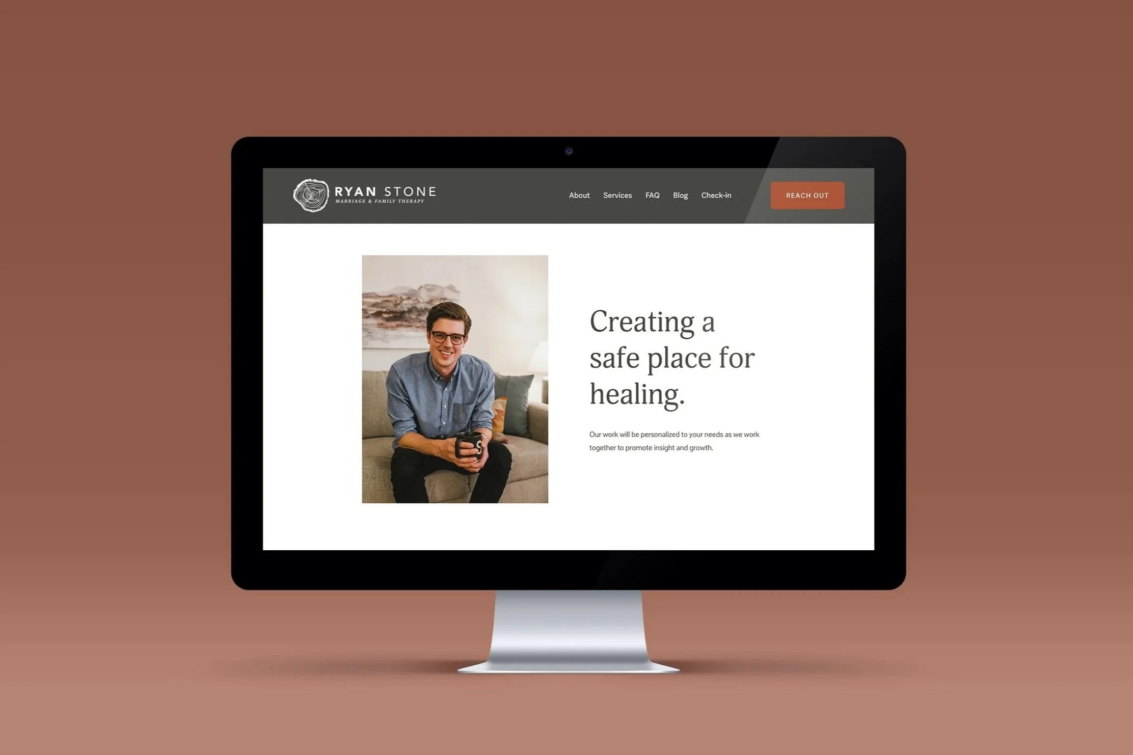 Computer monitor displaying a therapy website with a photo of a smiling man sitting on a couch holding a mug, and the text 'Creating a safe place for healing'.
