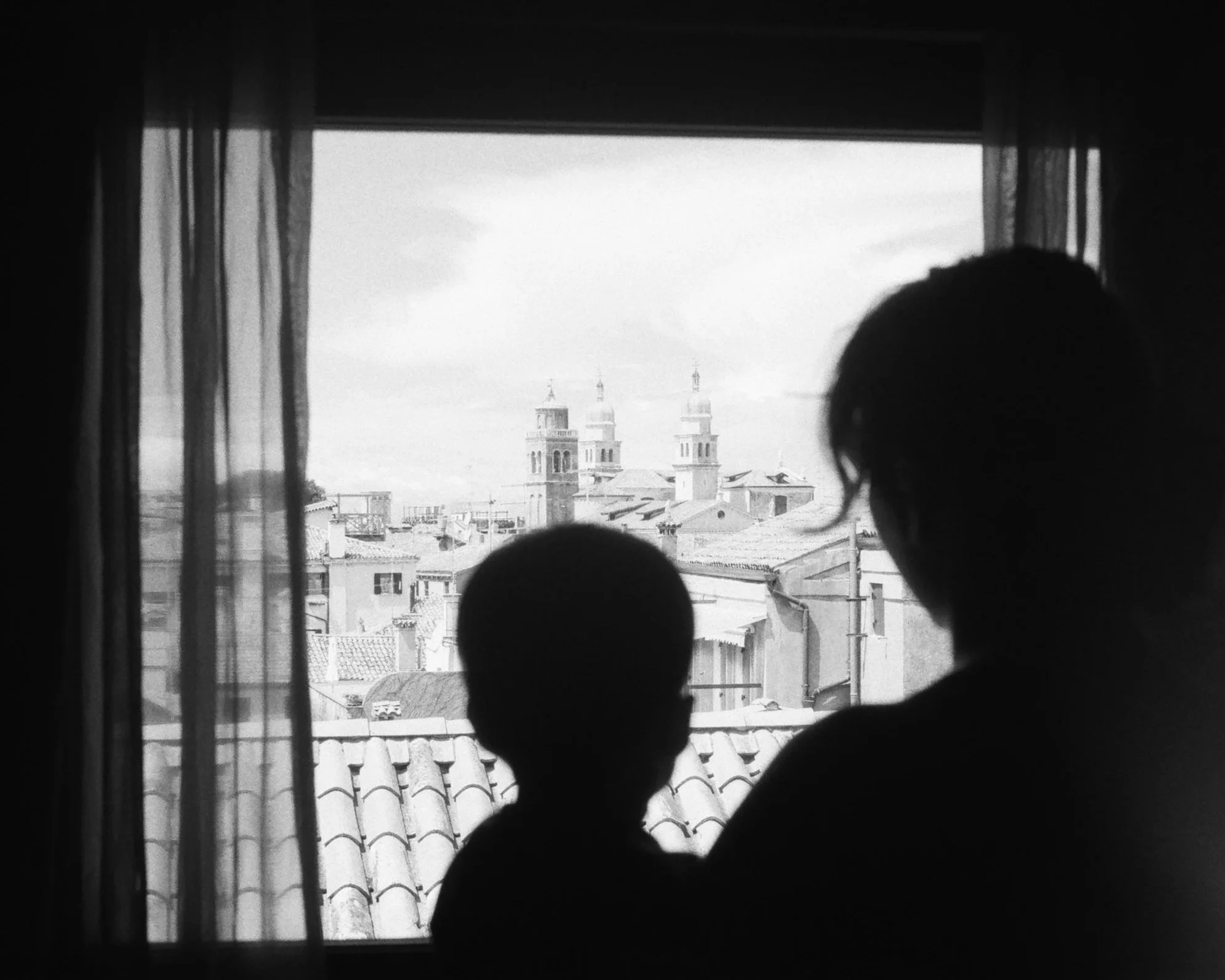 A black-and-white photo of a child and a person standing by a window with curtains, overlooking rooftops and church towers.
