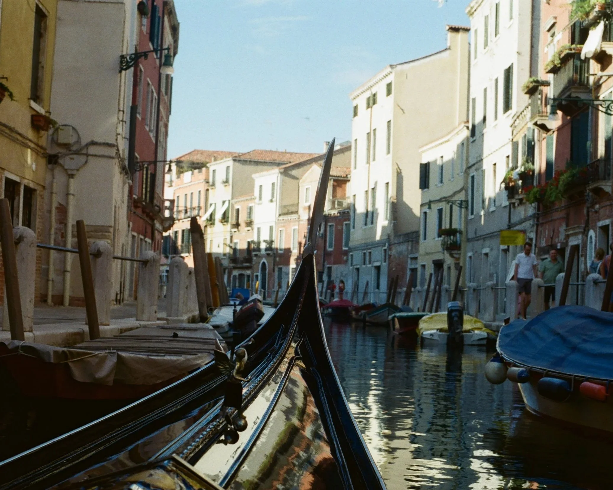 A canal with boats docked along the sides, in front of colorful buildings with windows and balconies, under a blue sky.