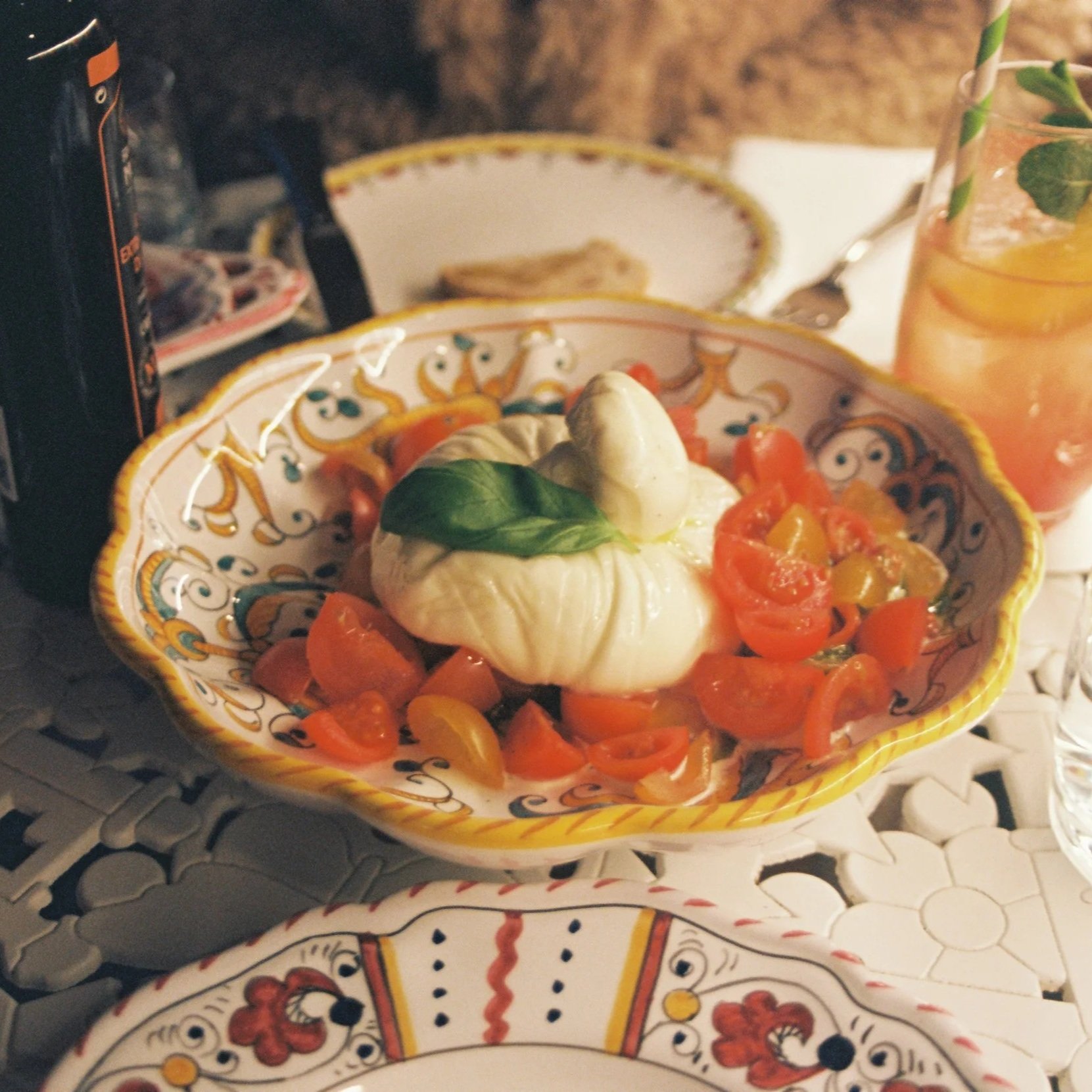 A decorative bowl filled with cherry tomatoes, topped with fresh mozzarella and basil leaves, on a table with a glass of iced beverage and a spoon nearby.