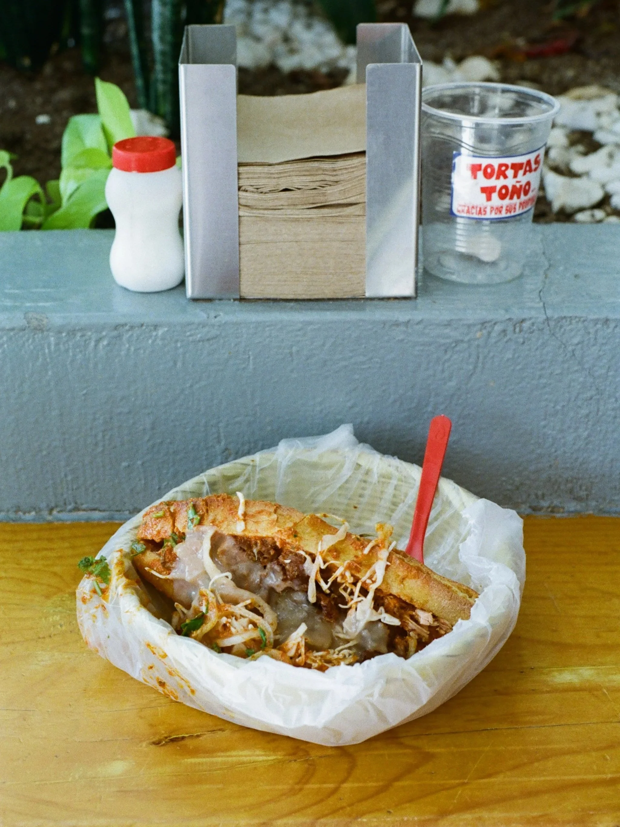 A half-eaten torta sandwich with meat, cheese, and vegetables in a paper wrapper on a wooden table. In the background, there are salt and pepper shakers, a napkin holder with paper napkins, and a clear plastic cup with a red and white label that read