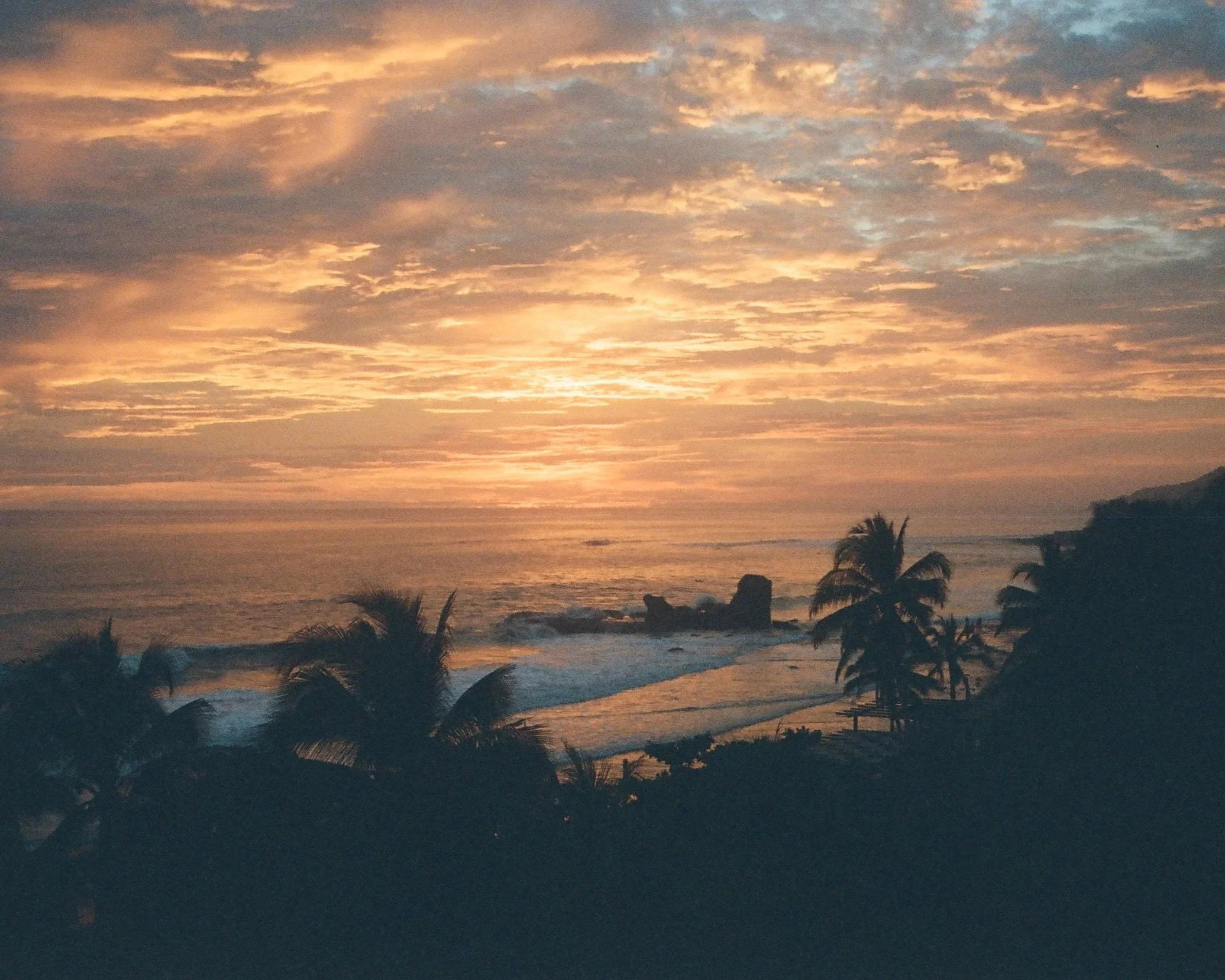 Sunset over the ocean with silhouetted palm trees in the foreground, partly cloudy sky with orange and pink hues.