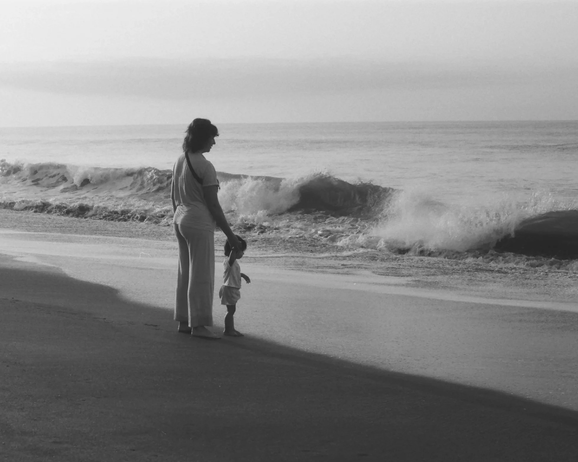 A woman and a young girl, presumably mother and daughter, stand on the beach near the shoreline, looking at the ocean waves as they crash onto the sand. The image is in black and white.