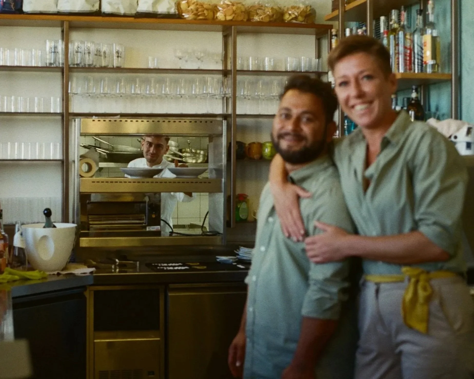 Two happy men are hugging in a kitchen or restaurant setting, with shelves of glasses, books, and kitchenware in the background. A chef is visible through a window behind them.