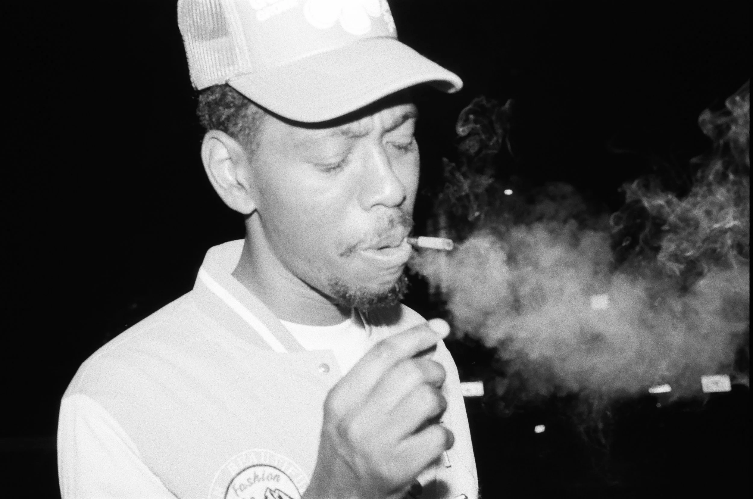 A man wearing a baseball cap and a shirt with a logo, smoking a cigarette and holding a small object, with smoke around him.