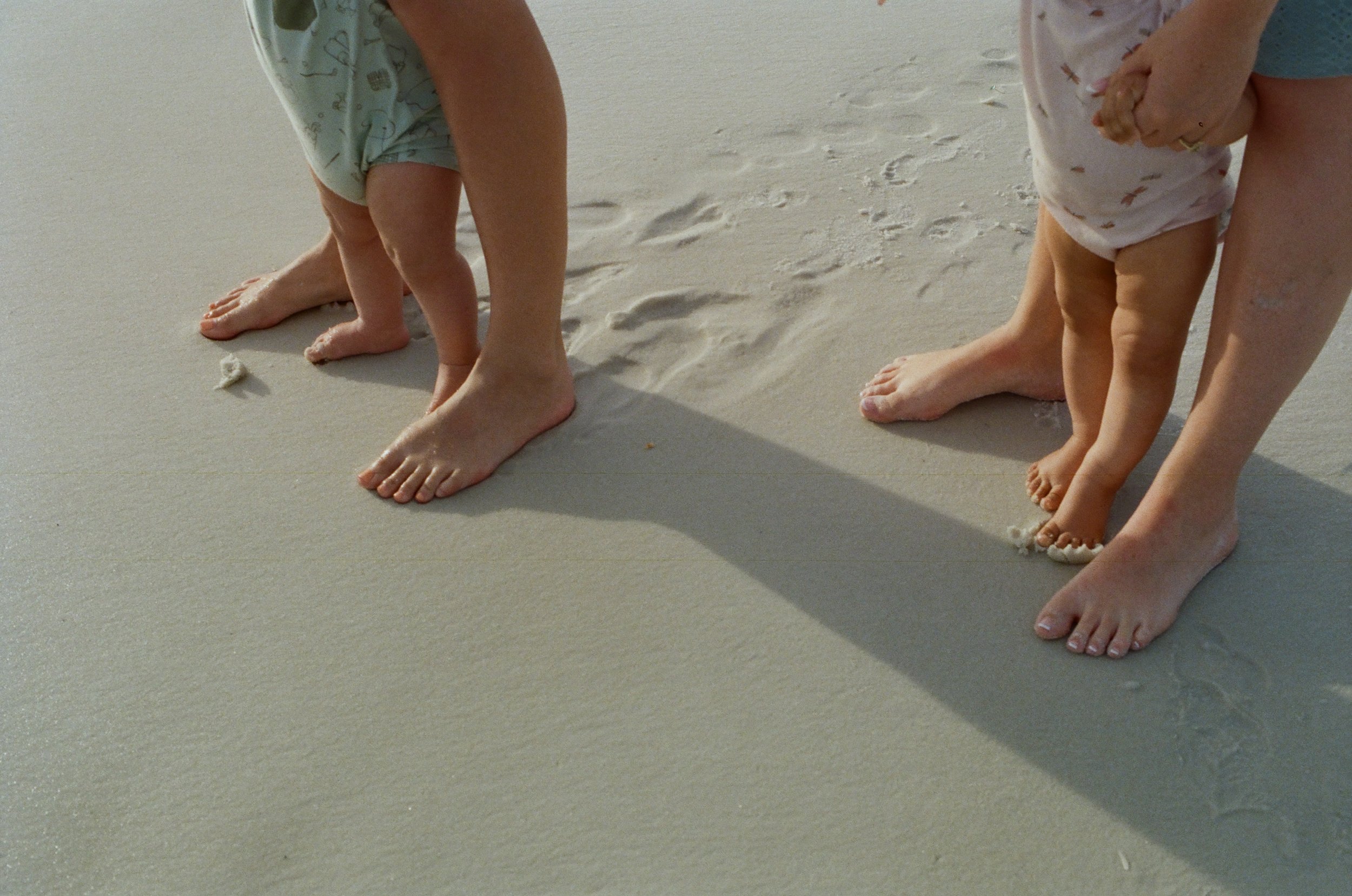 Close-up of a young child and an adult, both barefoot, bending over on sandy beach with footprints. The child is wearing a light-colored printed onesie, and the adult's leg is partially visible, wearing blue clothing.