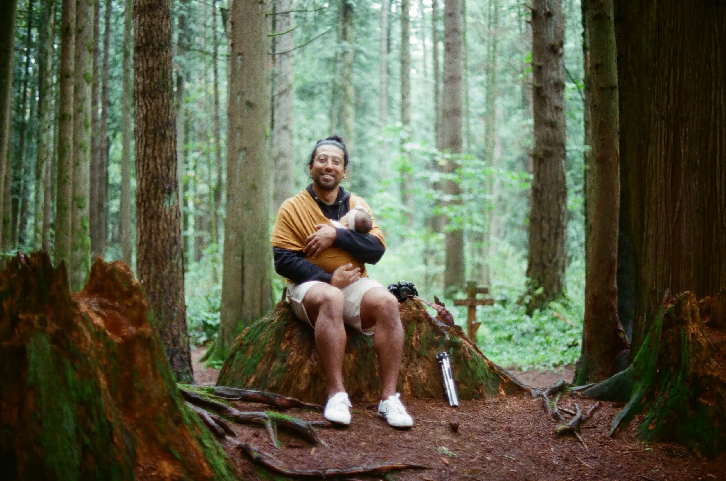 A man sitting on a tree stump in a dense forest holding a baby. He is smiling and wearing a yellow and black jacket, shorts, and white sneakers, with a camera on his lap and a walking cane beside him.