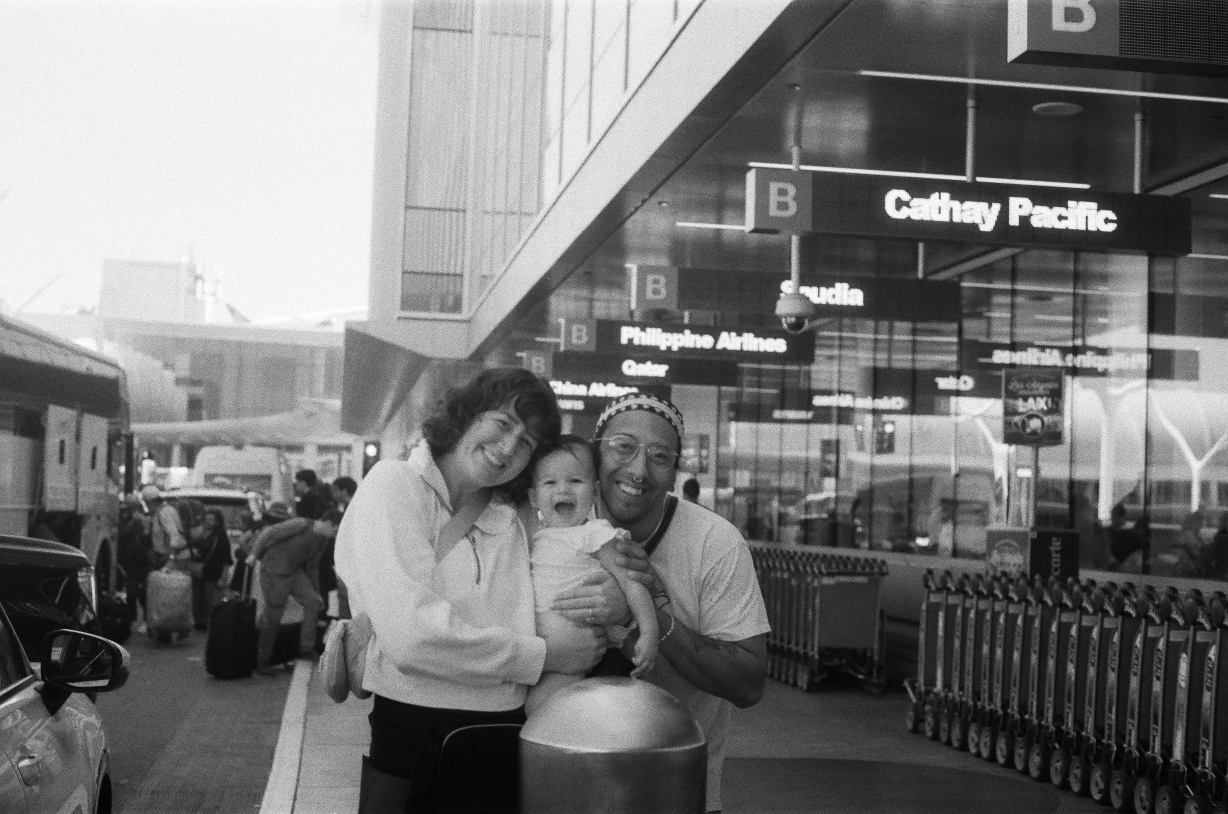 Four people, including a child, smiling and posing for a photo at an airport terminal near baggage claim. In the background, signs indicate different airlines and gates. The setting appears busy with airport activities and luggage.