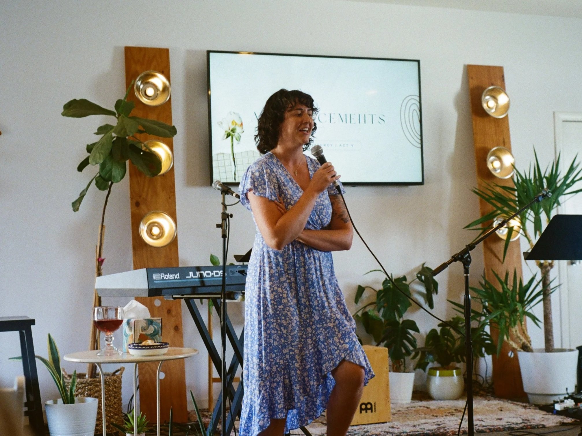 Woman in a blue floral dress speaking into a microphone during a presentation or event in a room with plants and decorative lighting.