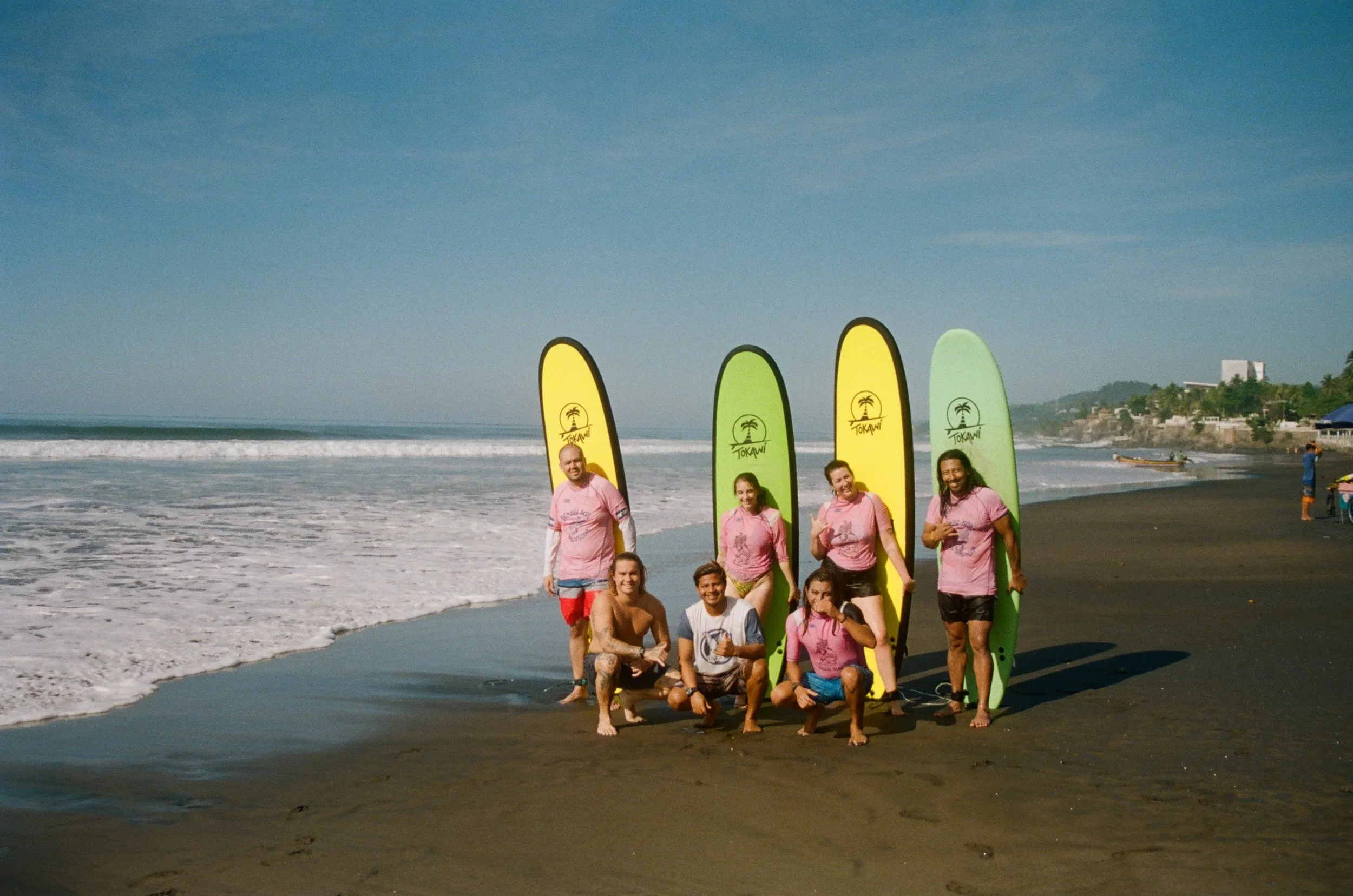 Group of eight people on a beach, smiling and posing for a photo with surfboards branded 'Tokawi' in the background, ocean waves and coastline visible.