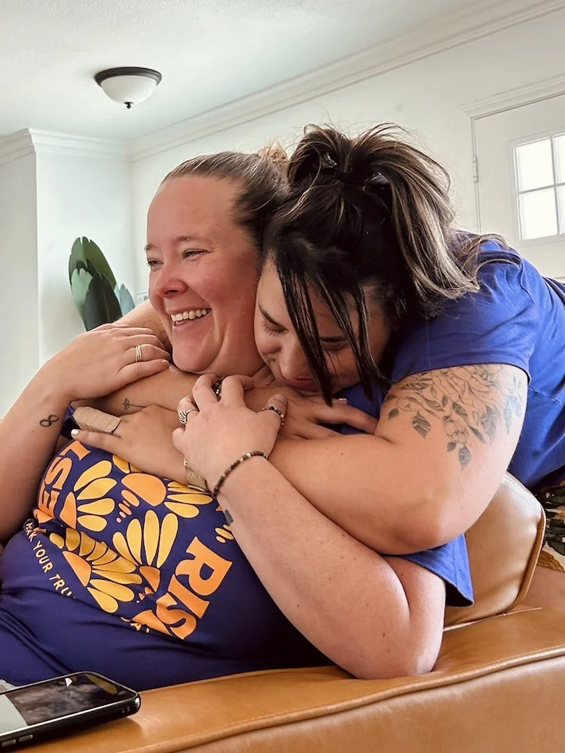 Two women sharing an affectionate hug, one smiling and the other with her face pressed into the first woman's shoulder, in a bright indoor setting.
