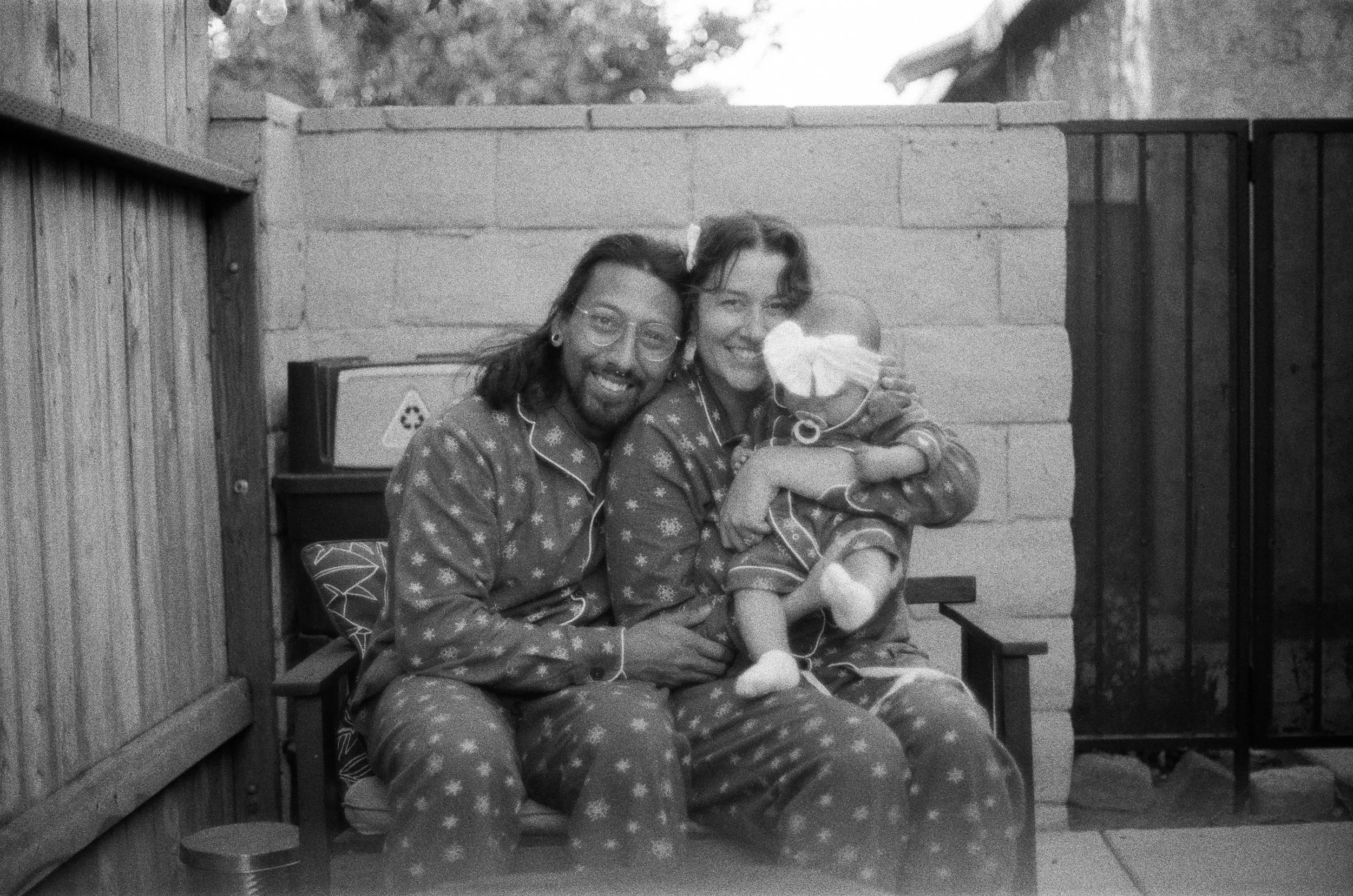 Two people in matching pajamas, a man with glasses and a woman, sitting outside on a bench and smiling, while the woman holds a small dog.