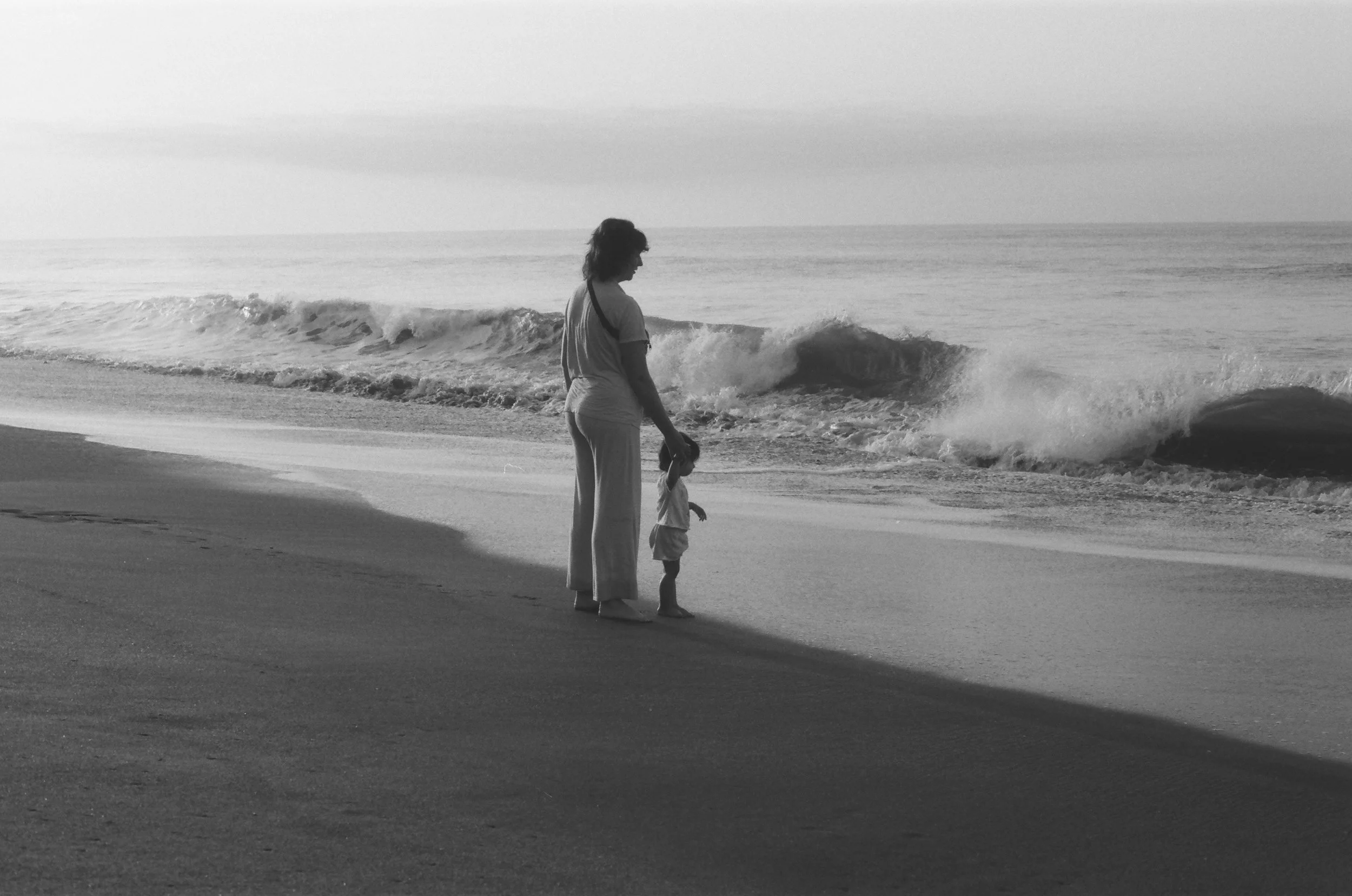 A woman and a child standing on the beach near the shoreline, watching the waves in black and white.