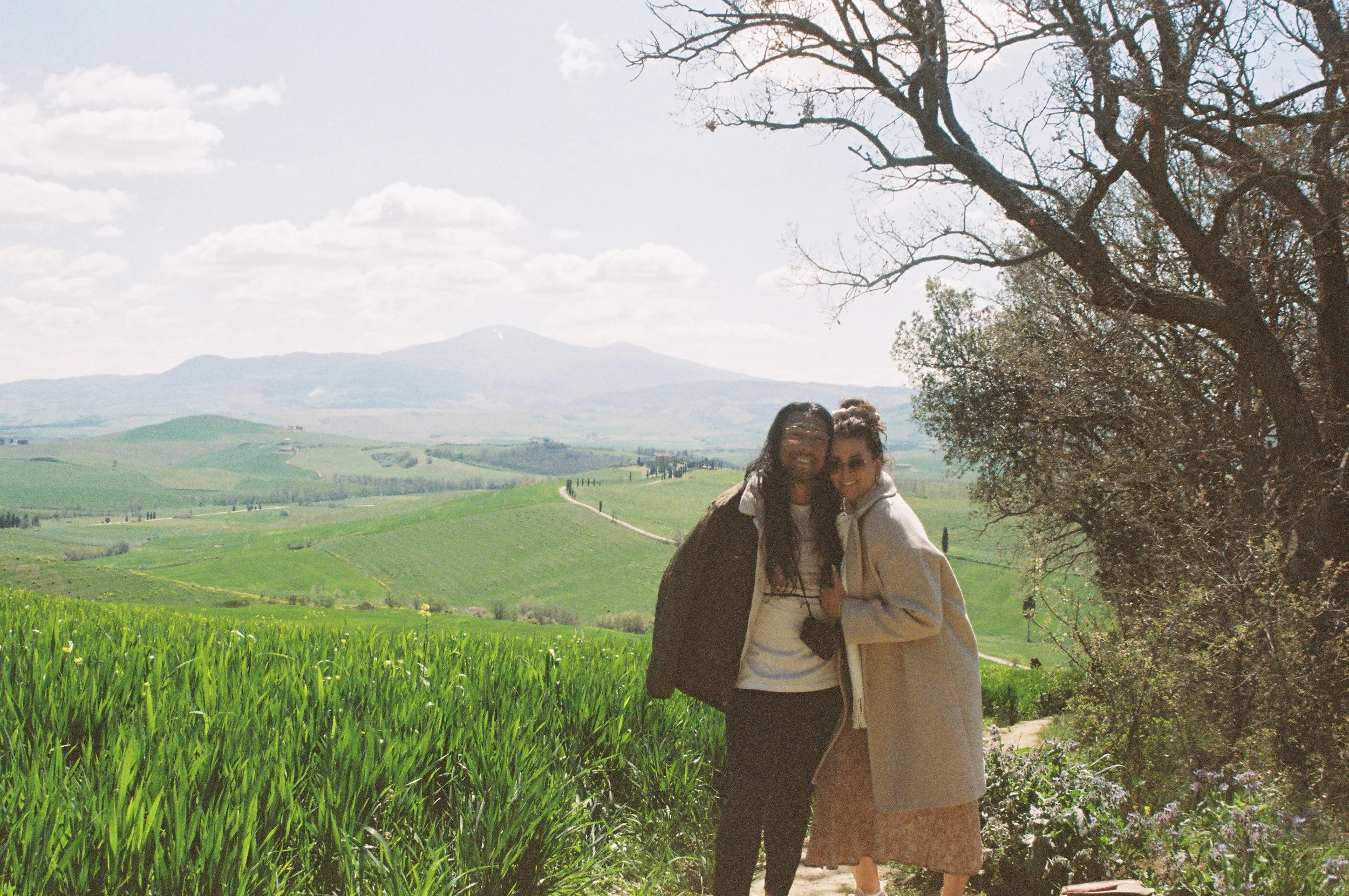 Two women smiling and hugging outdoors in a green, scenic landscape with rolling hills, mountain in the distance, and a large tree nearby.