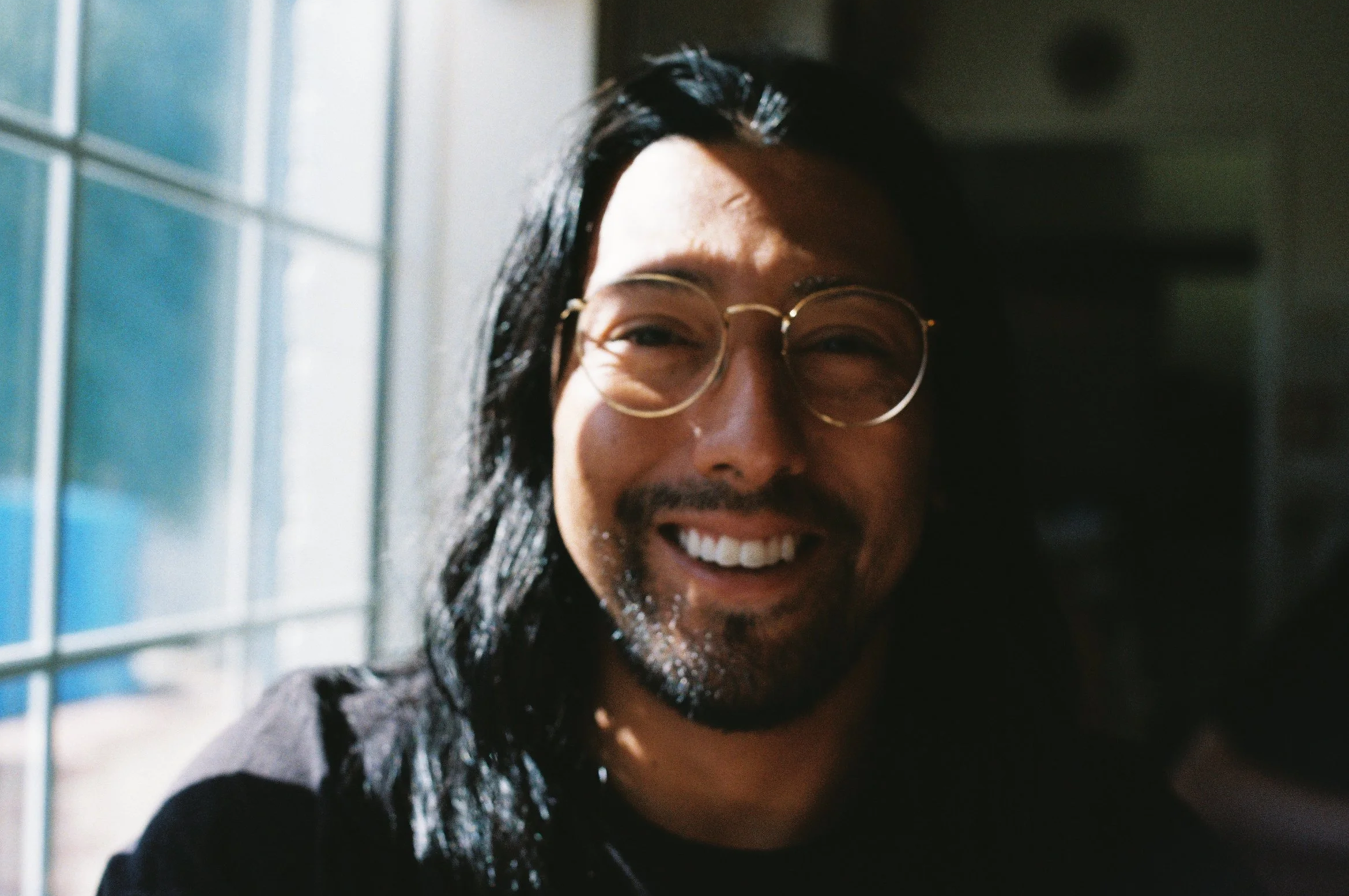 A person with long dark hair, glasses, and a beard smiling, standing near a window with sunlight.