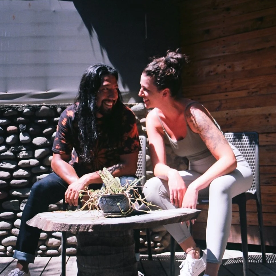 A man and woman sitting outdoors at a small wooden table, smiling and looking at each other. They are in a cozy setting with a stone wall and wooden paneling background, near a plant on the table.