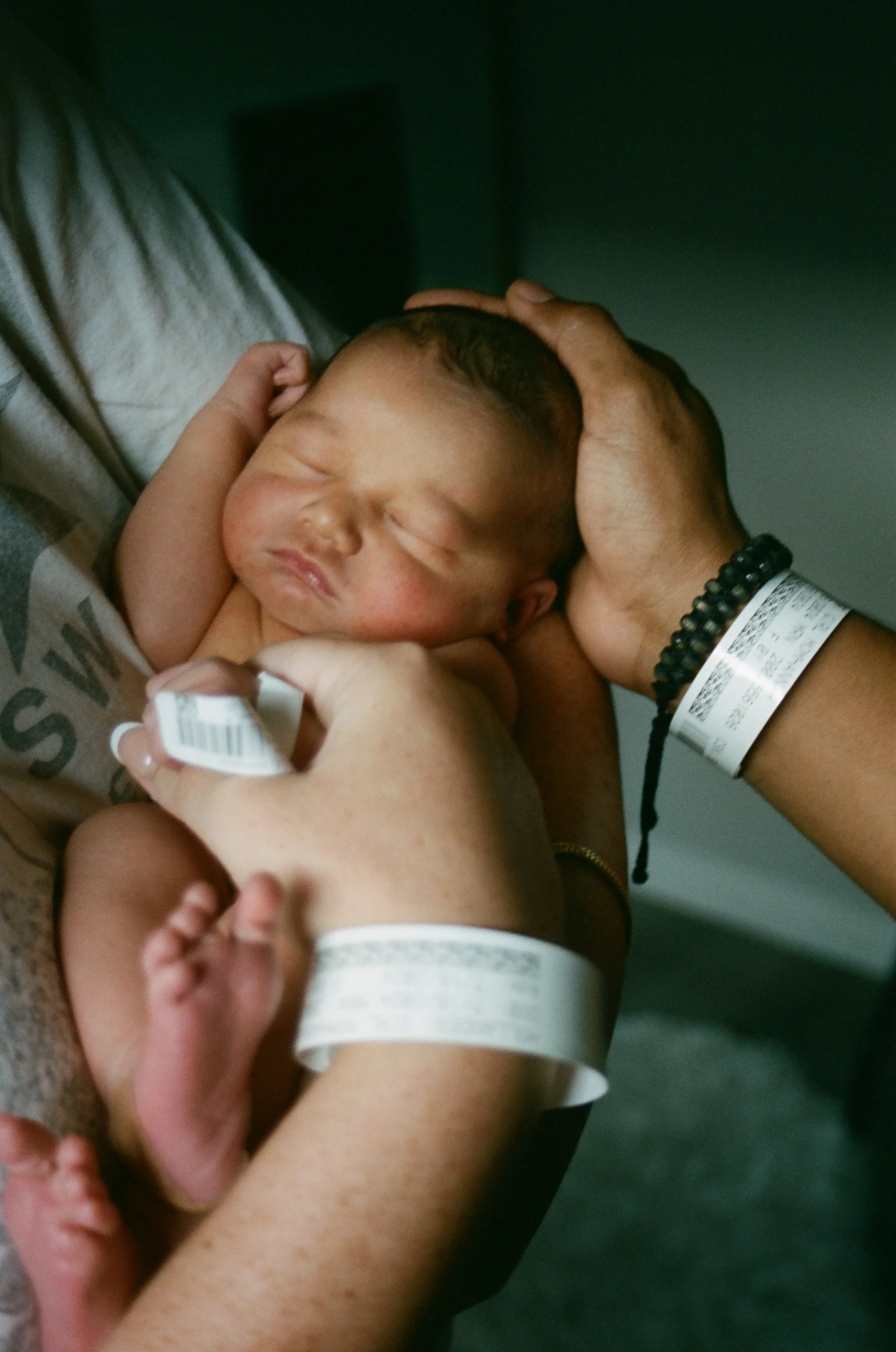 A newborn baby being held gently by multiple hands, with one hand supporting the baby's head and another holding the baby's body. The baby has closed eyes and a peaceful expression, with hospital wristbands visible on the hands holding the baby.