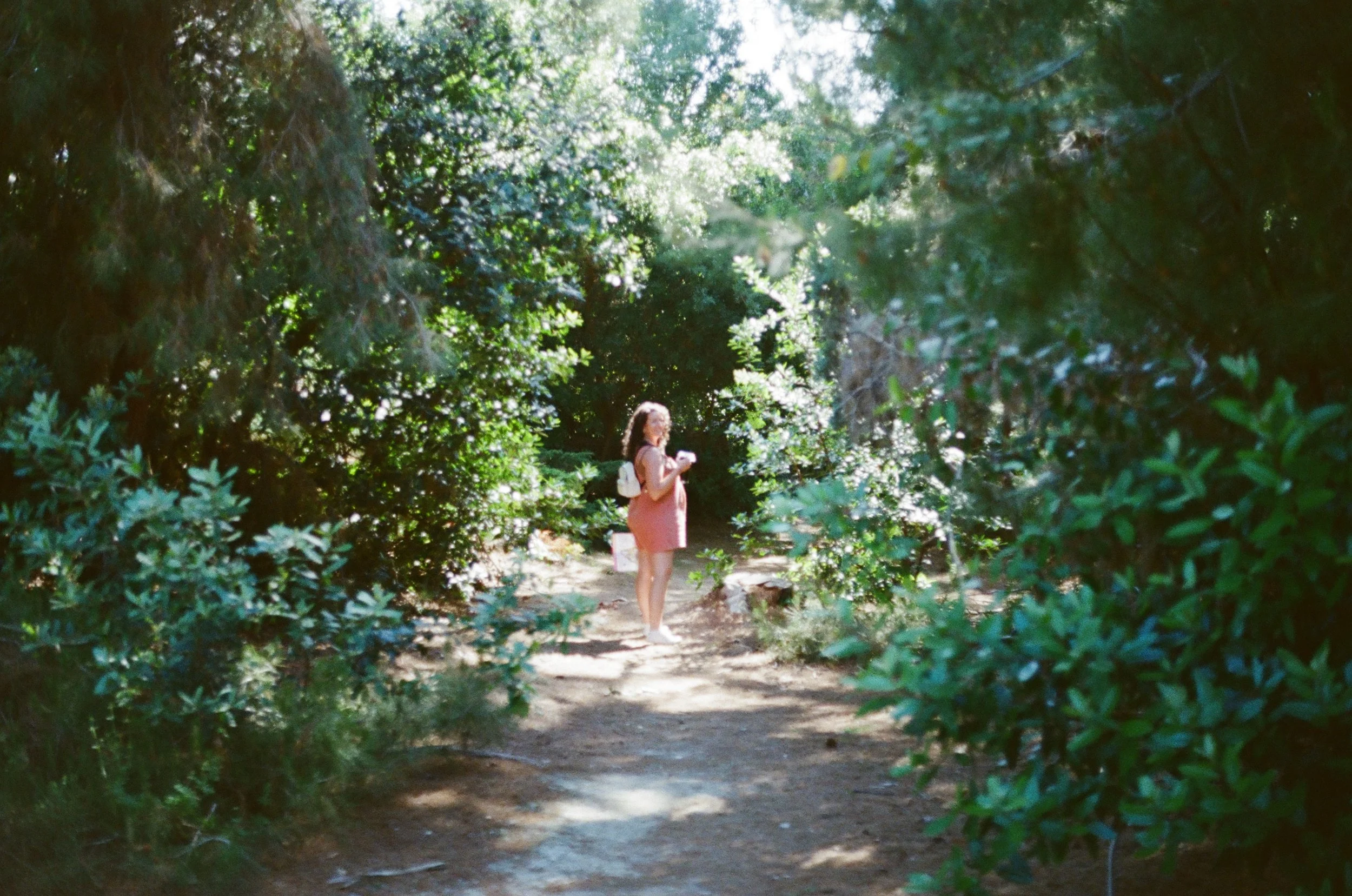 A woman standing on a forest trail, surrounded by lush green trees with sunlight filtering through the leaves.
