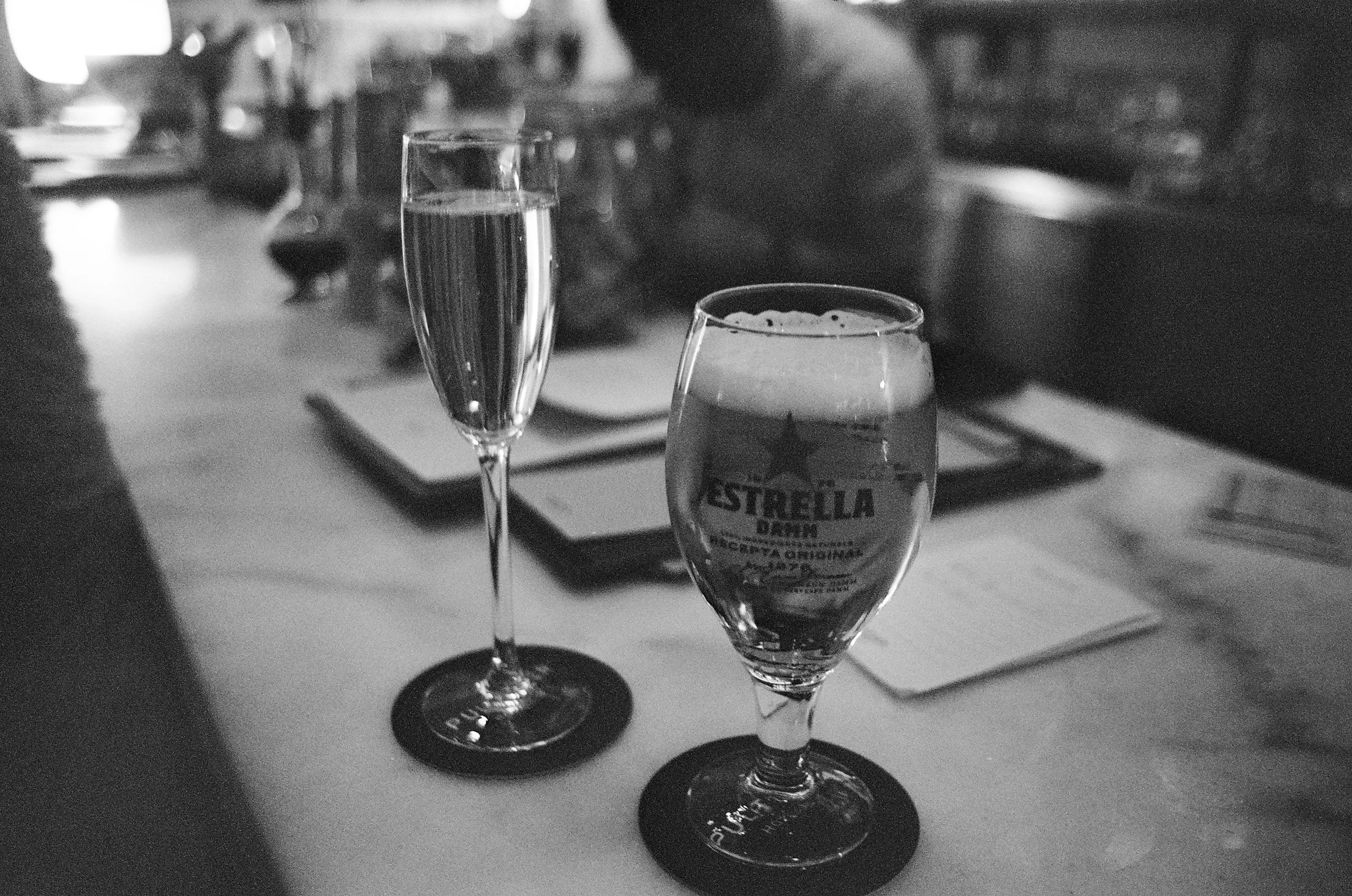 Two filled champagne flutes on a restaurant table, with one glass labeled Stella D'oro. Out of focus in the background are plates and a tray, suggesting a dining setting.
