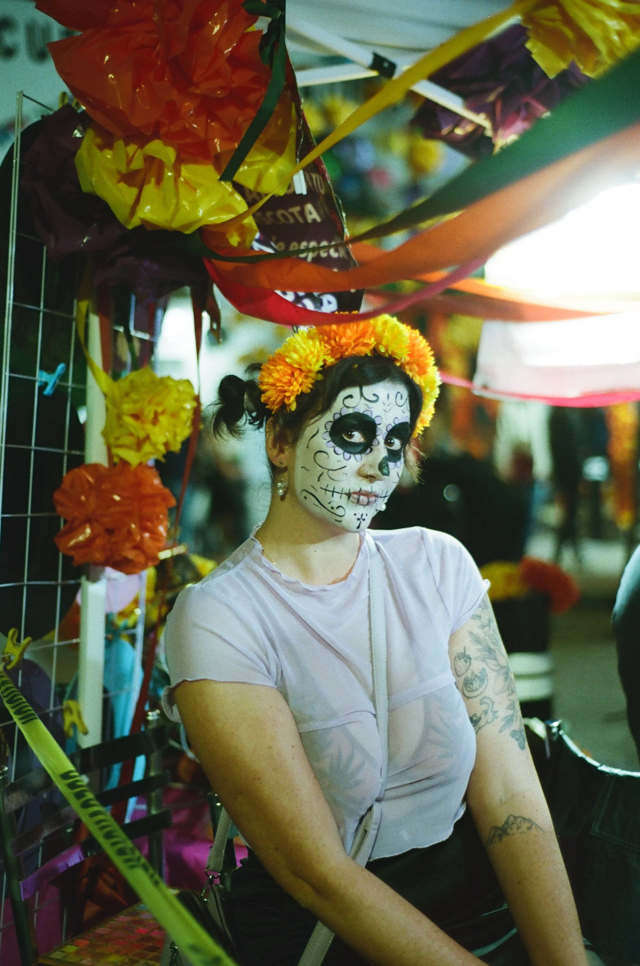 A woman with tattoos and dark hair in pigtails wearing white face makeup with day of the dead sugar skull design, a floral crown, and a light pink shirt, sitting in front of colorful items and decorations at a festive event or market.