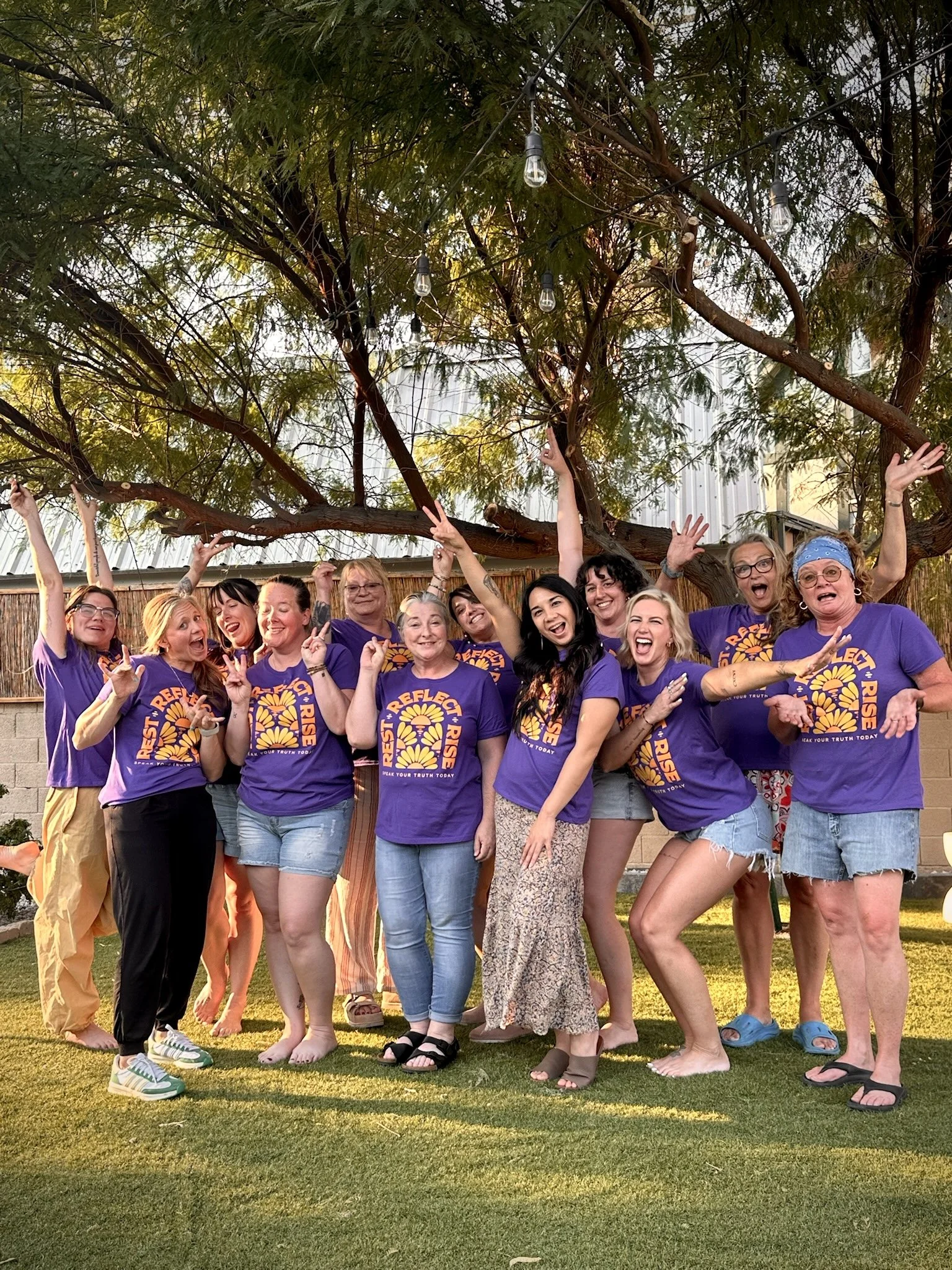 A group of women wearing purple T-shirts with a logo and text on the front, standing outdoors on grass under tree branches with string lights. They are smiling, making gestures, and seem happy.