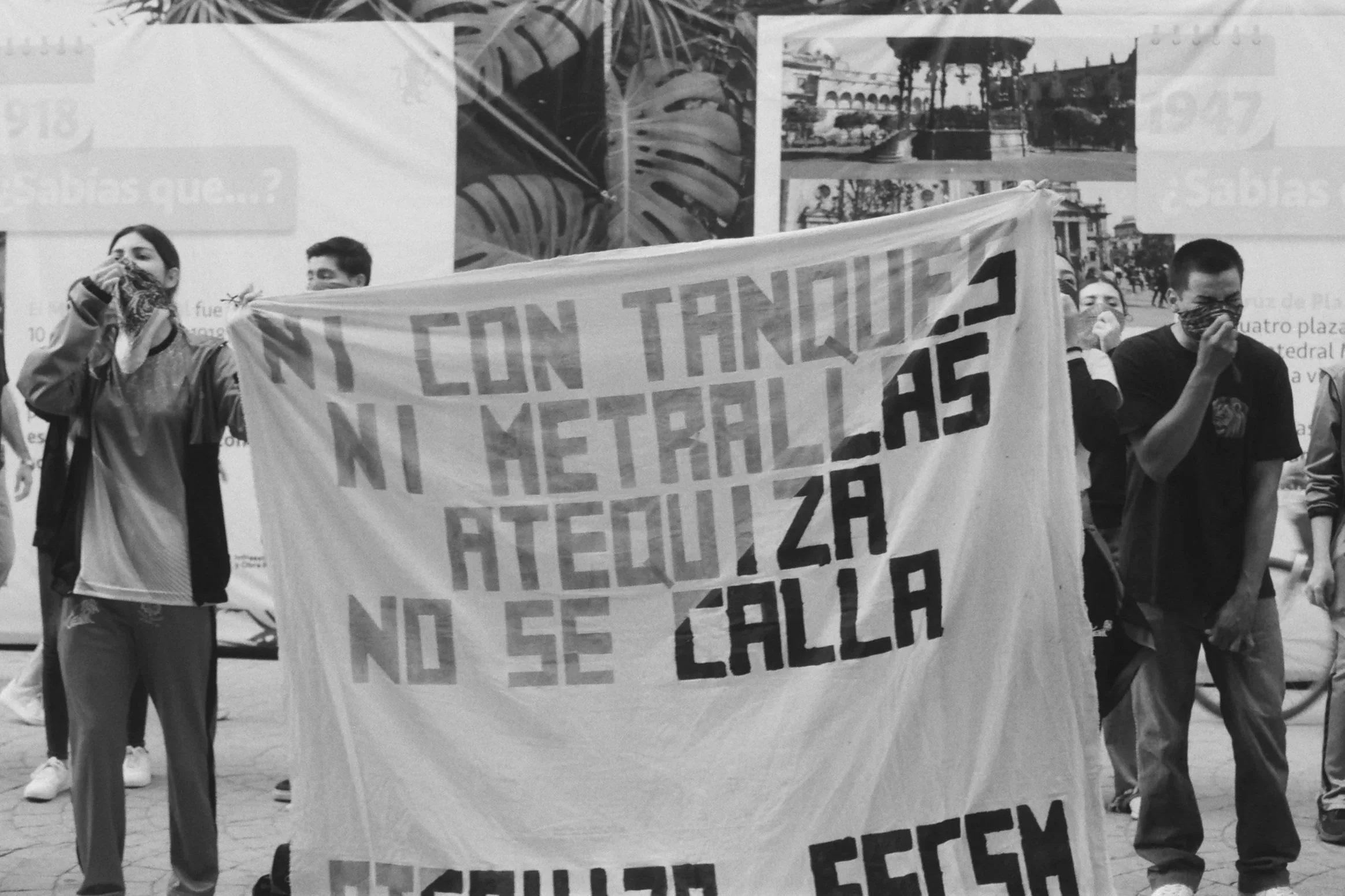 People participating in a protest march on a city street, holding a large banner with printed text. Facemasks worn by some participants, background includes architectural landmarks and banners.