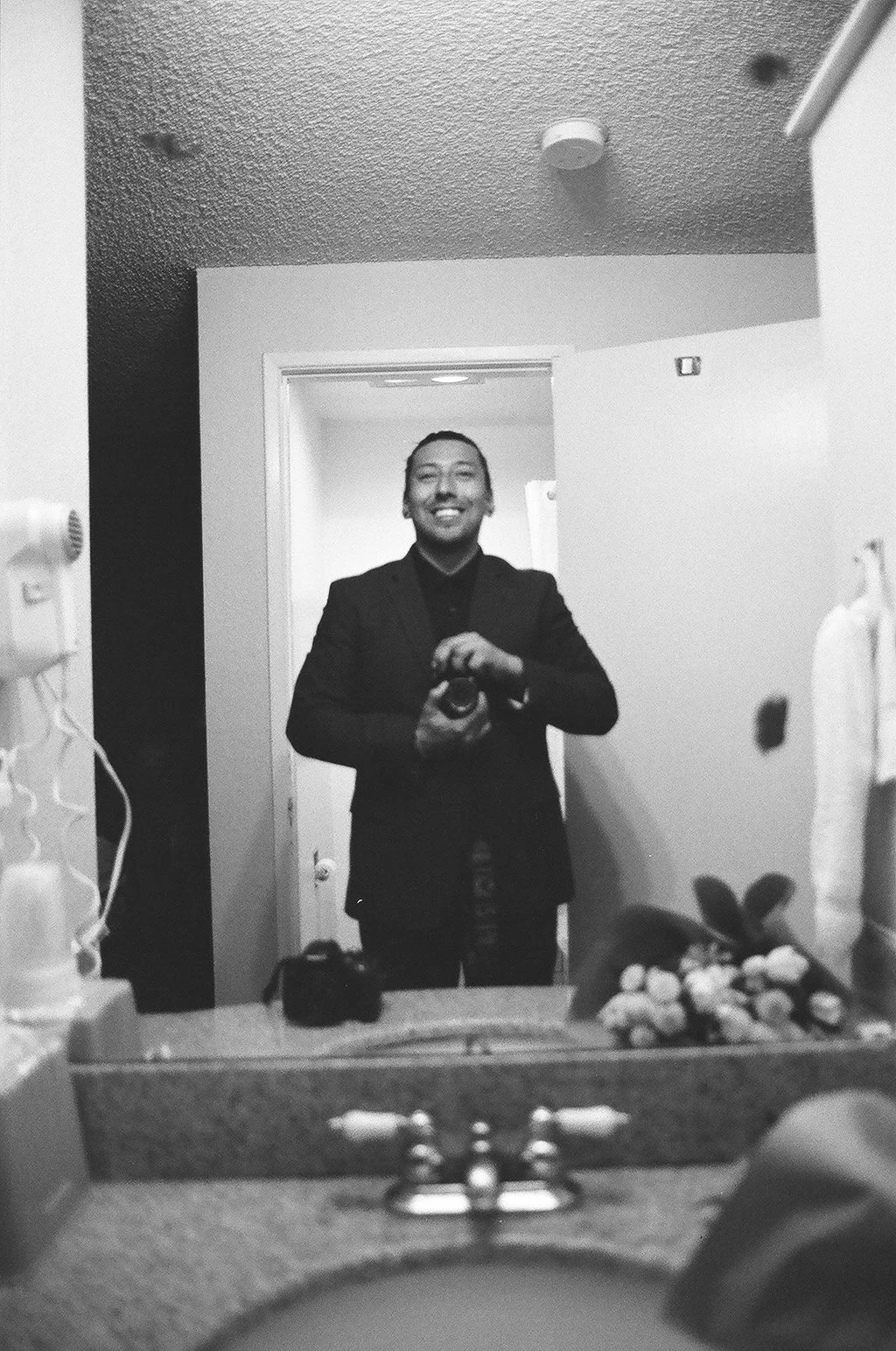 A man in a suit smiling and taking a photo of himself in a mirror, in a room with a bathroom sink and various items around.
