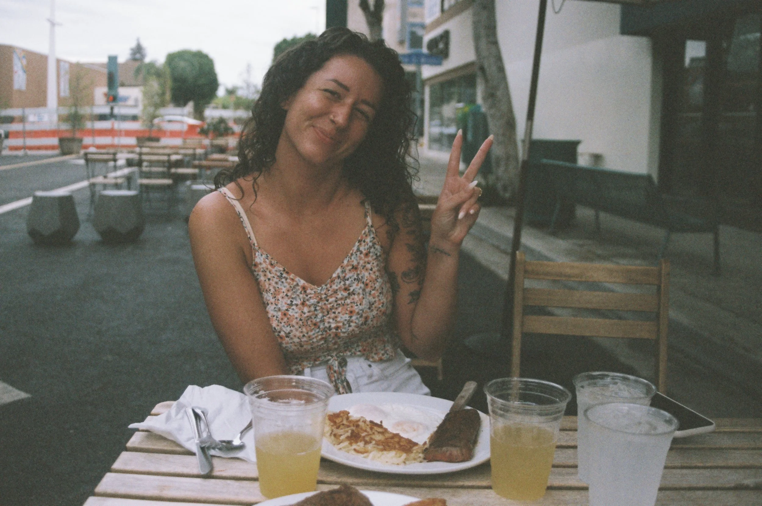 Woman with dark curly hair making a peace sign and smiling while sitting outdoors at a table with pizza, drinks, and food containers, with a background of an outdoor seating area and street.