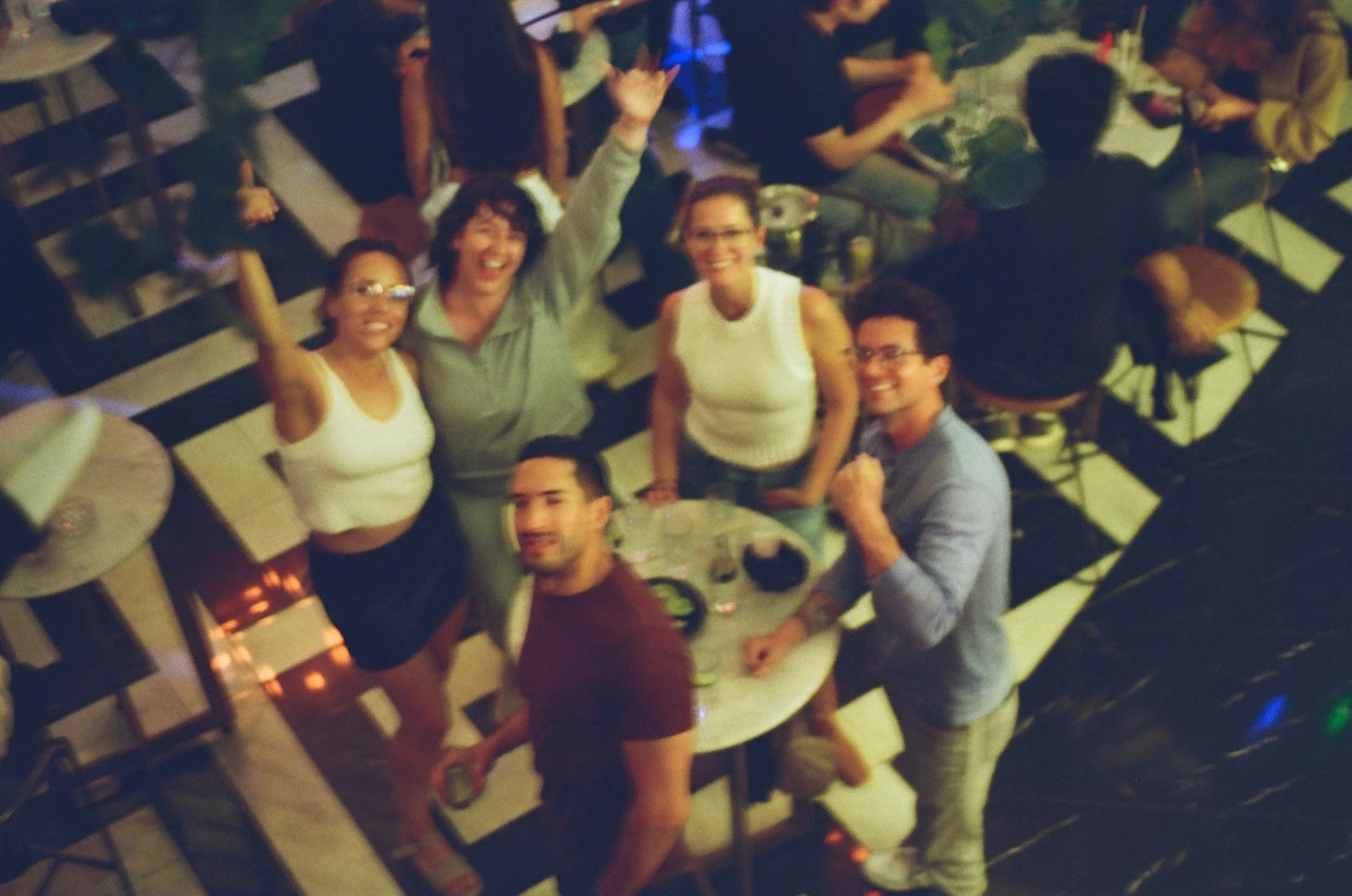 A group of people at a party or celebration, gathered around a table with drinks. Some are smiling and looking up at the camera, which is held above them. The atmosphere appears lively and joyful.
