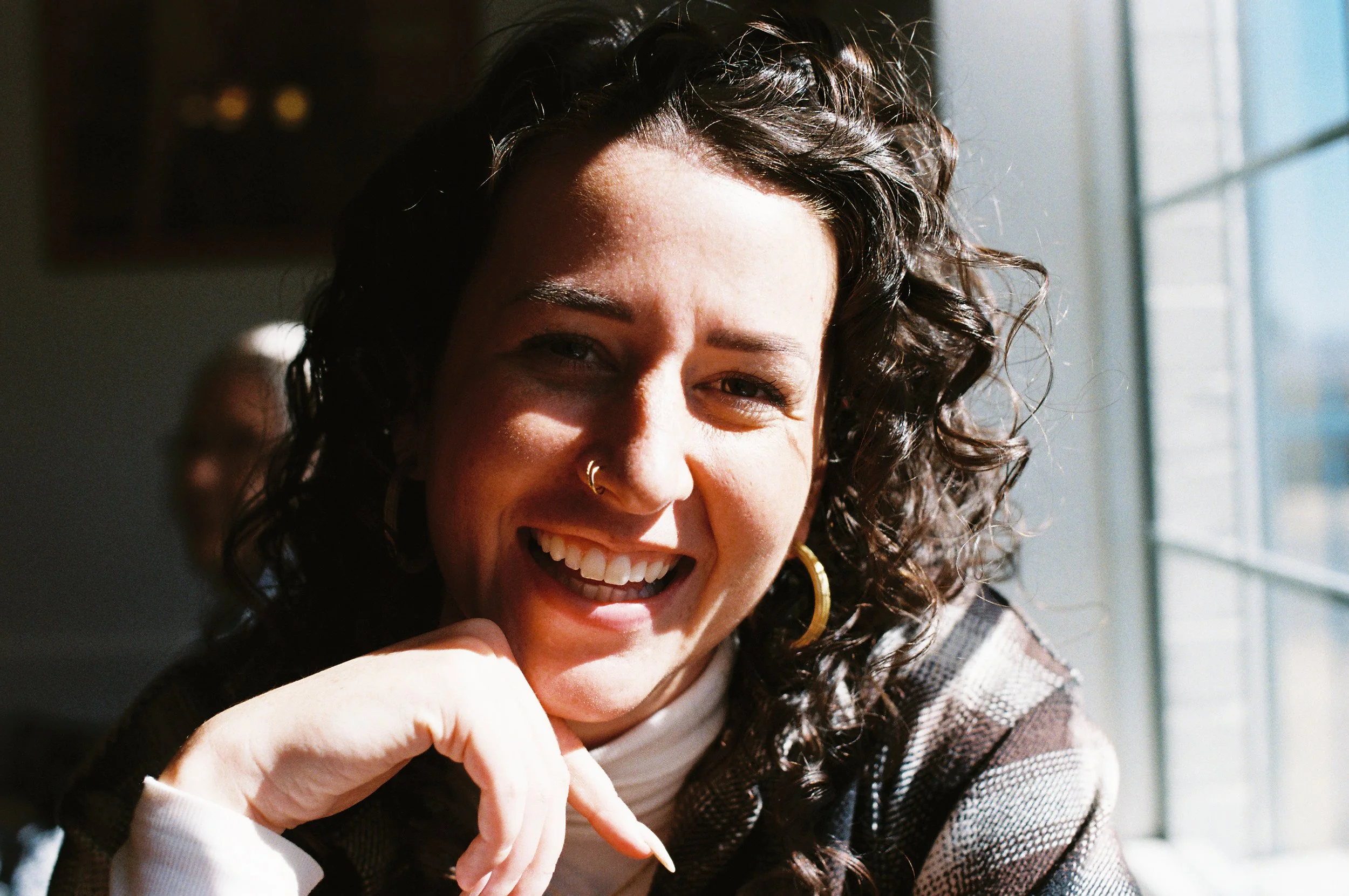 Close-up of a woman with curly hair, smiling, with sunlight streaming through a window behind her.