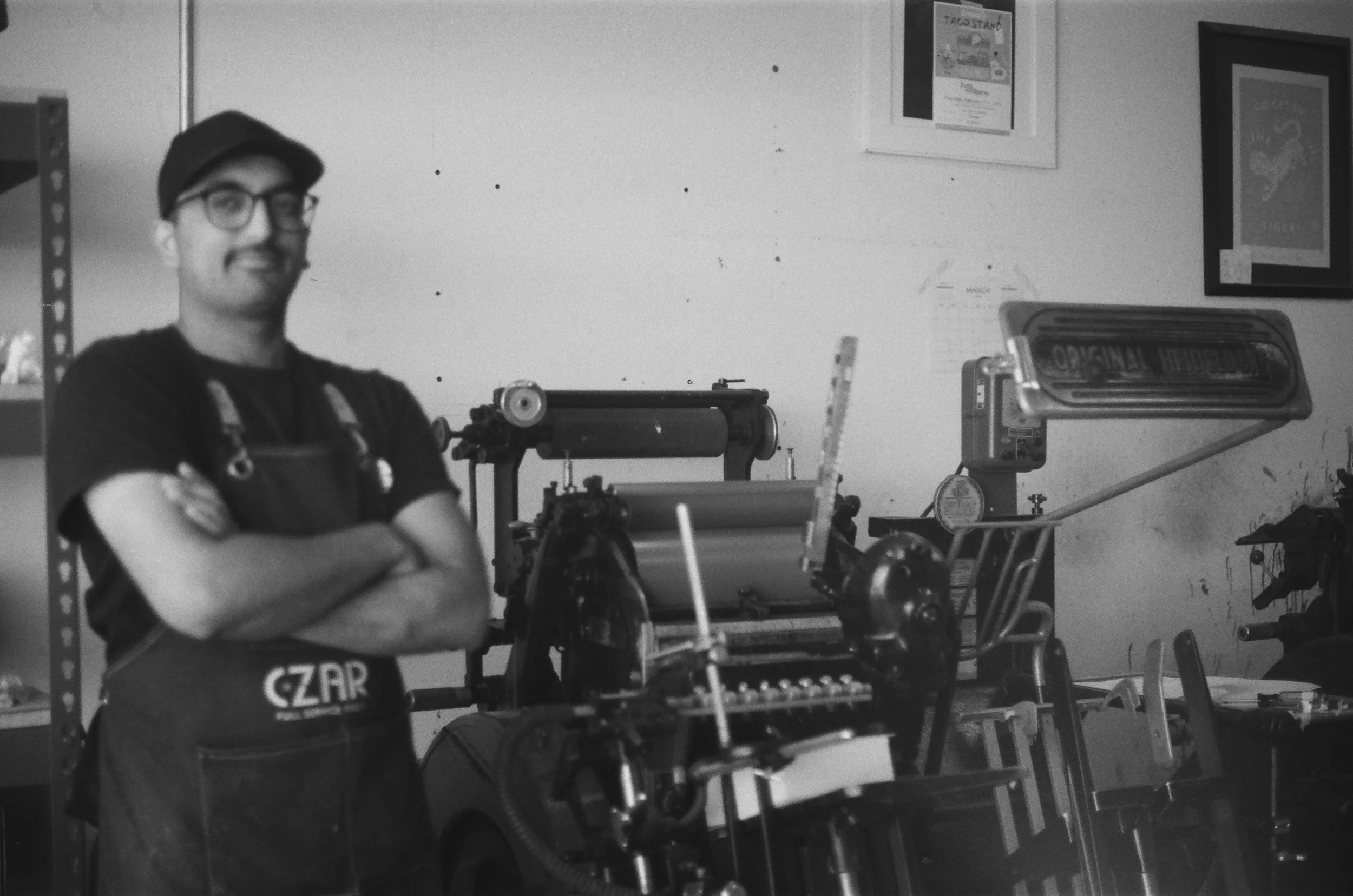 A man wearing glasses, a cap, and an apron standing with his arms crossed in a workshop surrounded by machinery and tools.
