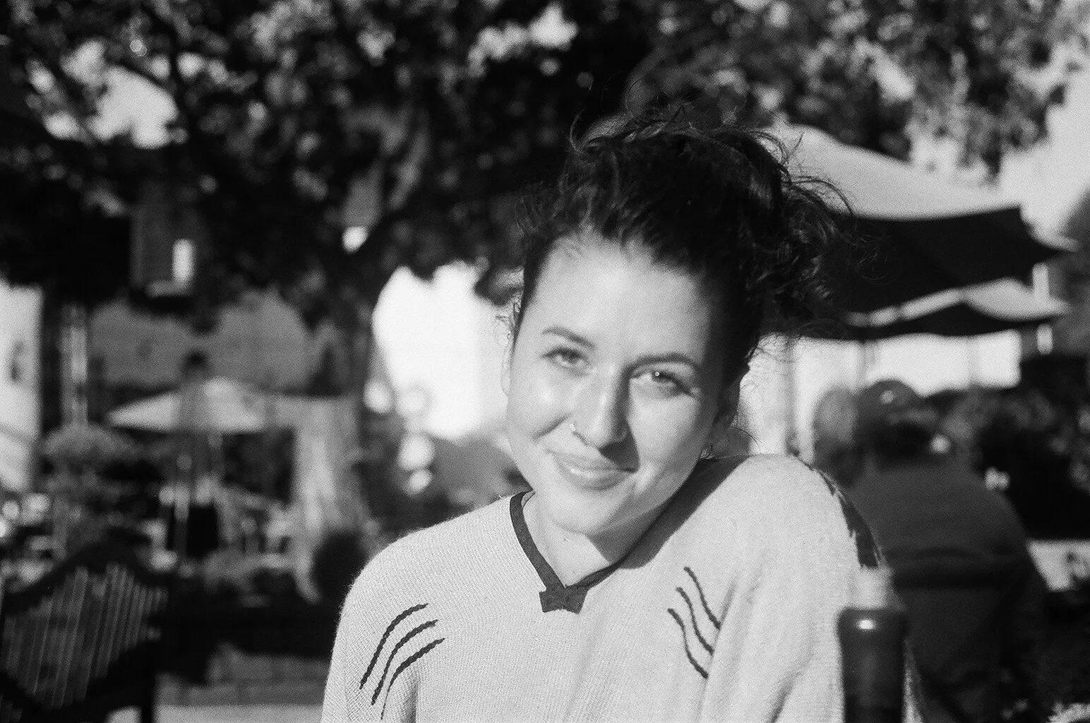 A woman with curly hair smiles at the camera while sitting outdoors, with trees and outdoor restaurant umbrellas in the background.