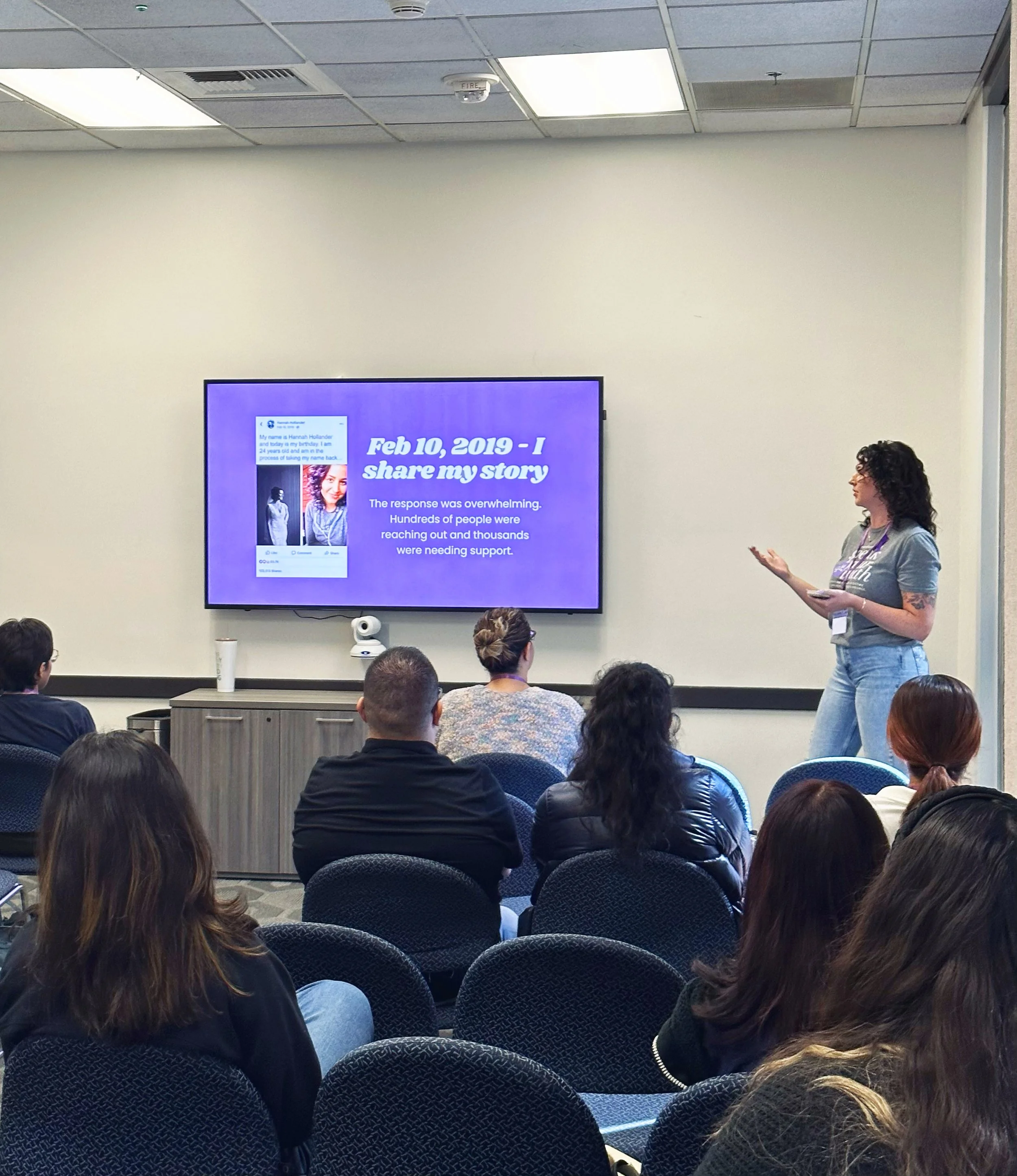 A woman giving a presentation in a conference room filled with seated attendees. A large screen behind her displays a slide with text about sharing a personal story on February 10, 2019.