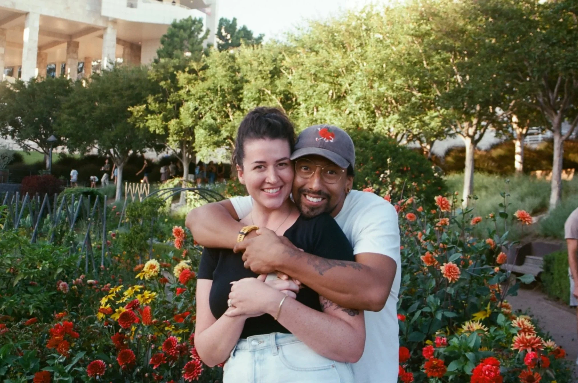 A happy couple hugging in a garden filled with orange and yellow flowers, with trees and a building in the background.