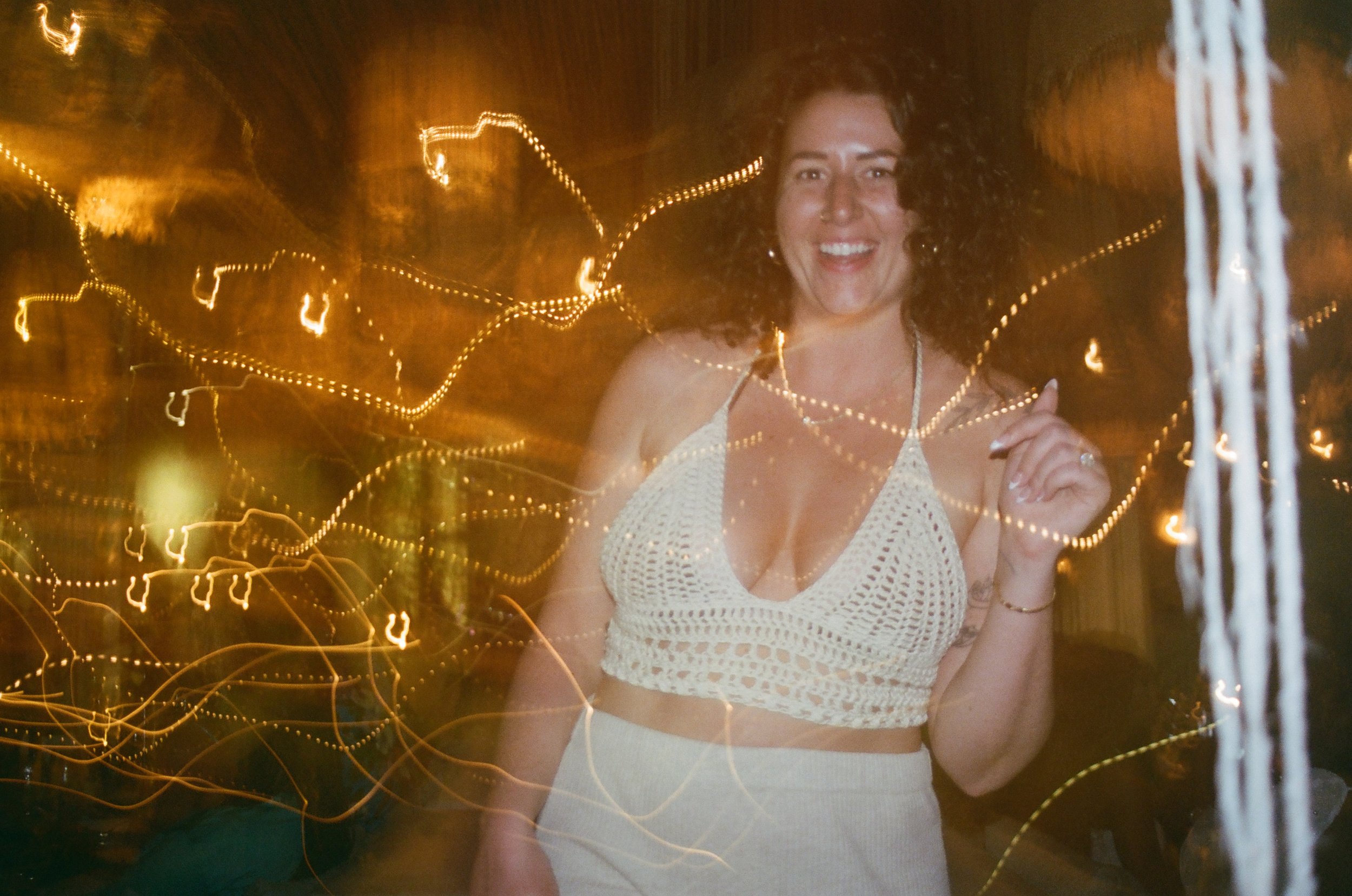 A smiling woman with curly dark hair, wearing a white crochet top and matching skirt, standing in front of string lights at night.