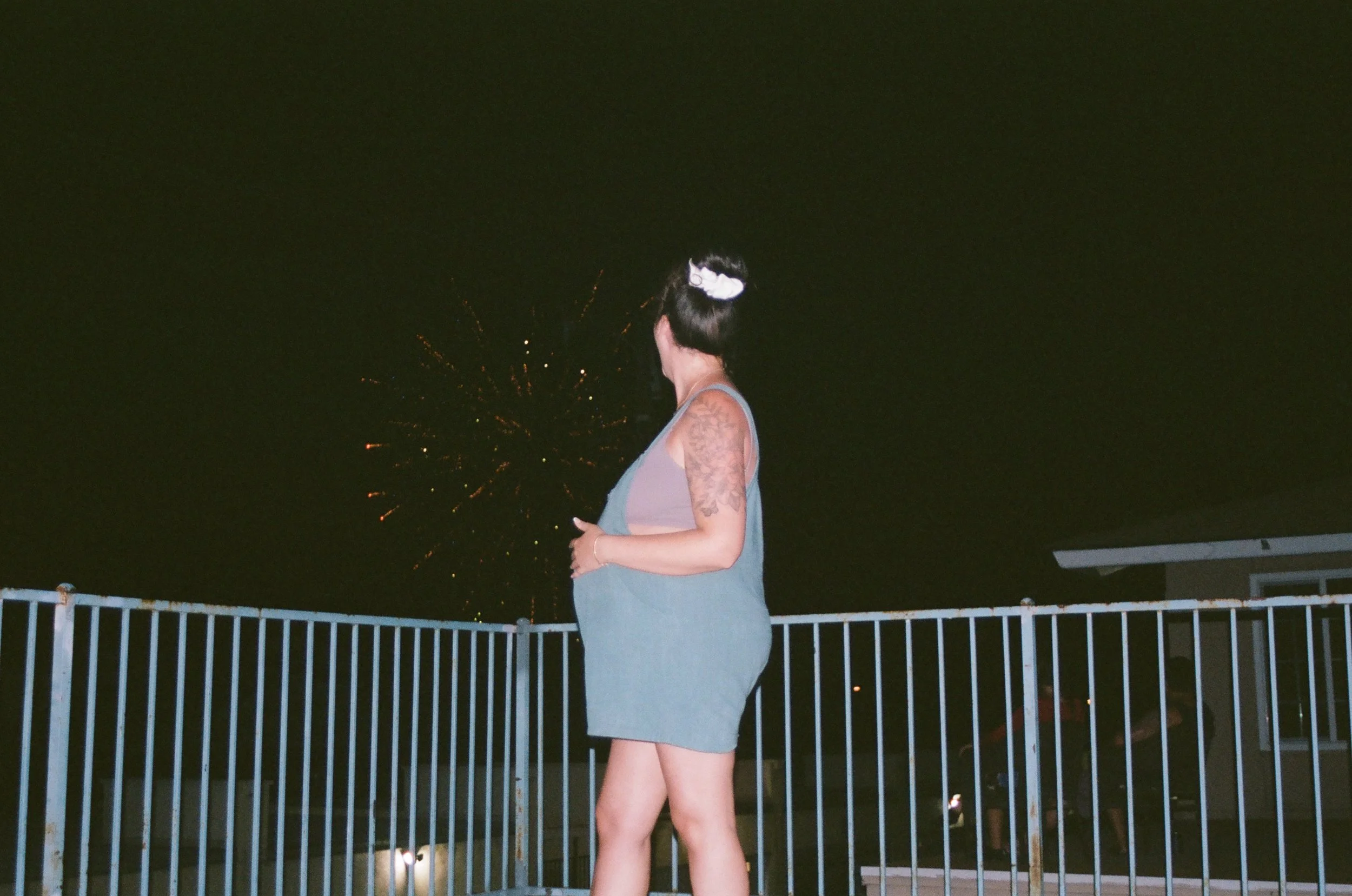 A woman with black hair tied up, wearing a gray sleeveless dress and tattoos on her arms, standing on a balcony at night with fireworks in the sky behind her.
