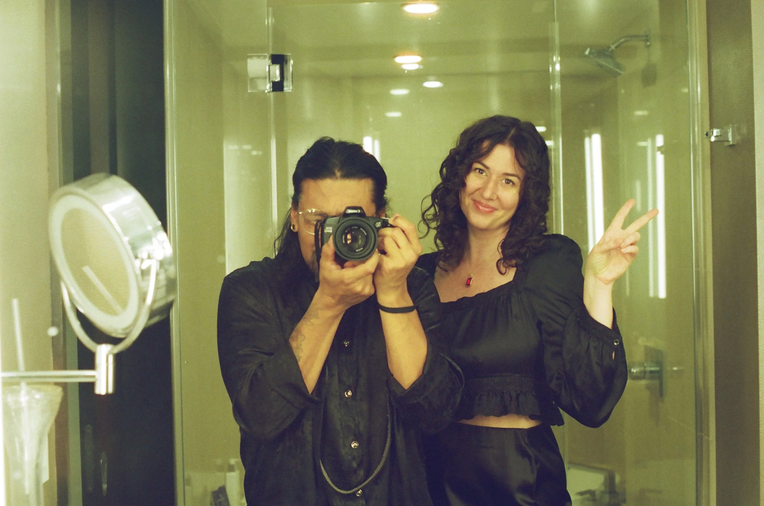 Two women taking a mirror selfie in a bathroom. One woman is holding a camera and the other is flashing a peace sign and smiling.
