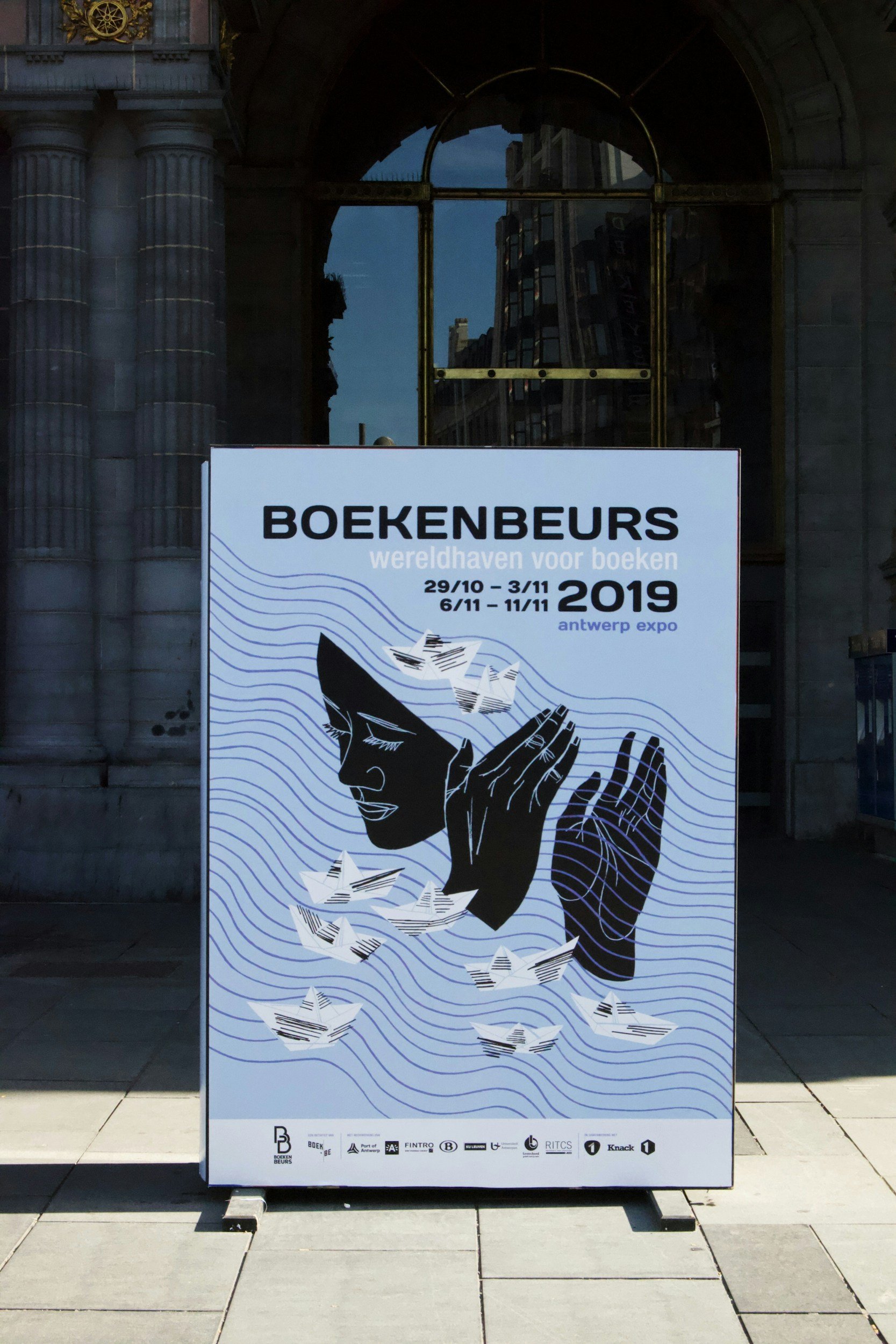 A white sign with blue lines and black text promotes the Book Fair at the Antwerp Expo in 2019, with illustrations of a woman reading and paper airplanes.
