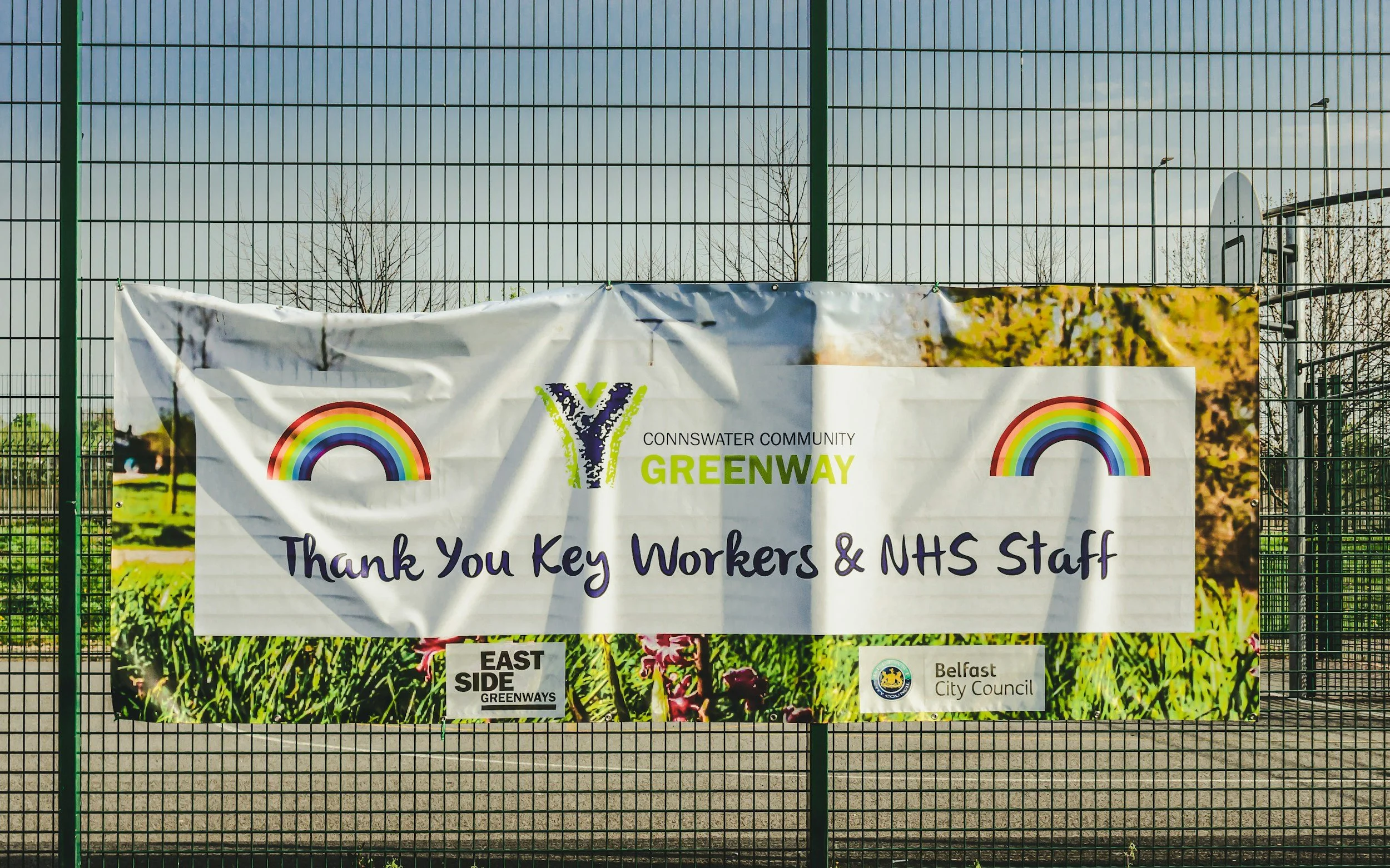 Banner on a green metal fence thanking key workers and NHS staff, with logos for Belfast City Council and East Side Greenways, featuring images of nature and rainbows.