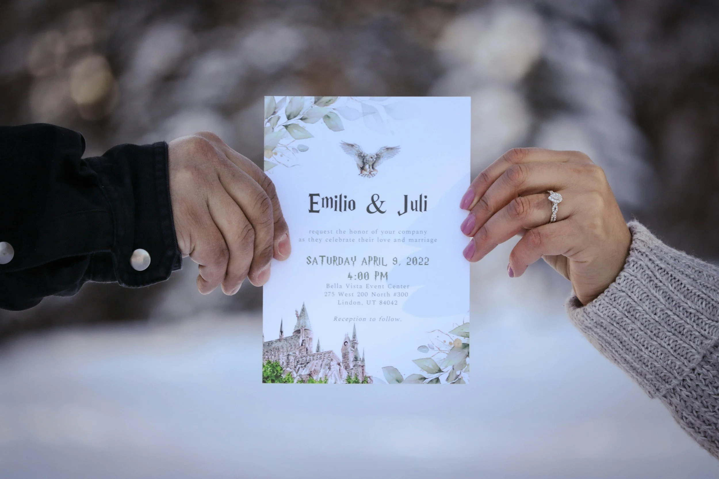 Two hands holding a wedding invitation, one hand with a wedding ring. The invitation has floral and castle illustrations, and details about a wedding date and location.