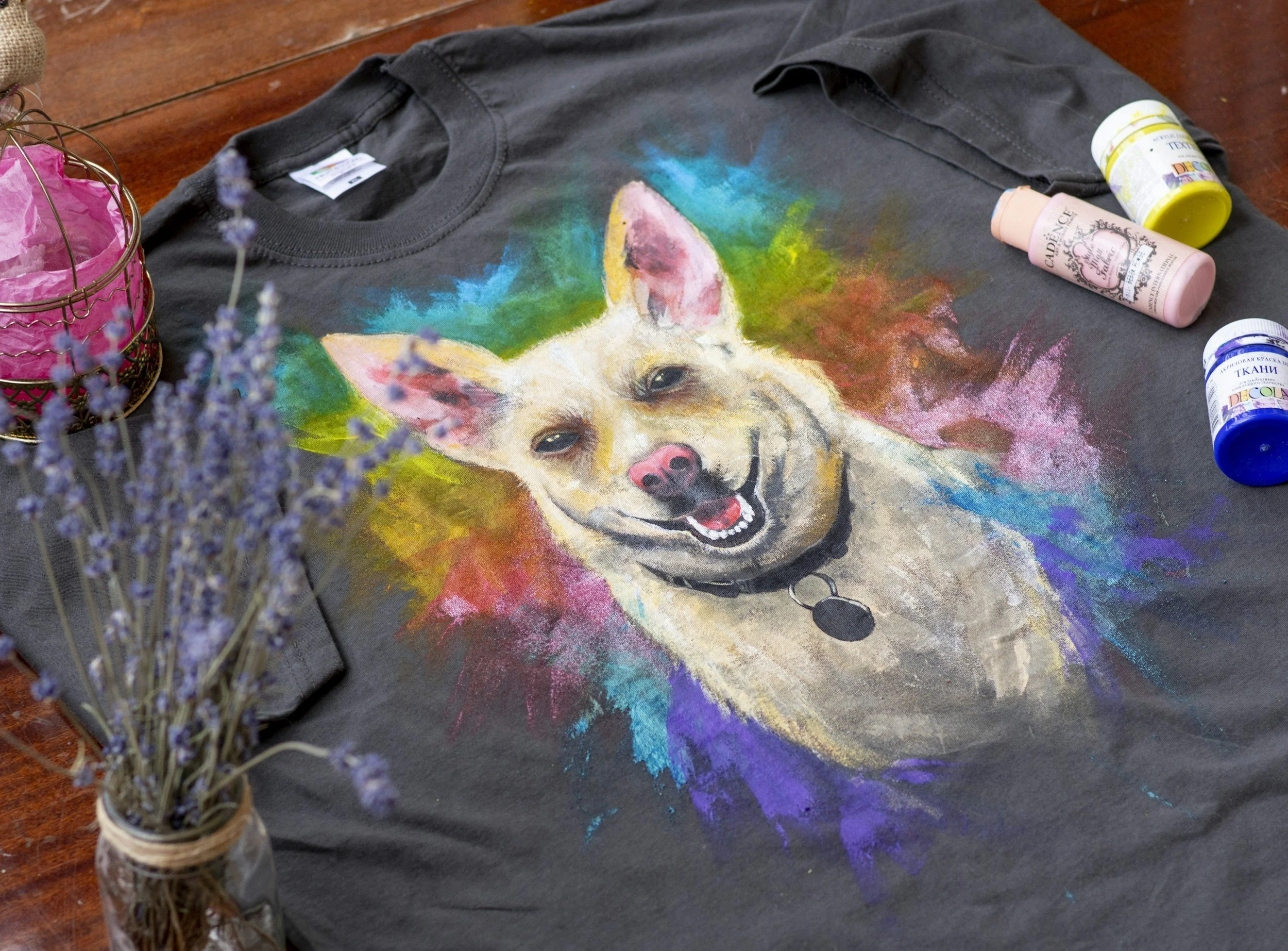 A black t-shirt lying flat on a wooden surface, featuring a colorful, artistic portrait of a smiling dog with its tongue out, surrounded by vibrant splashes of color.
