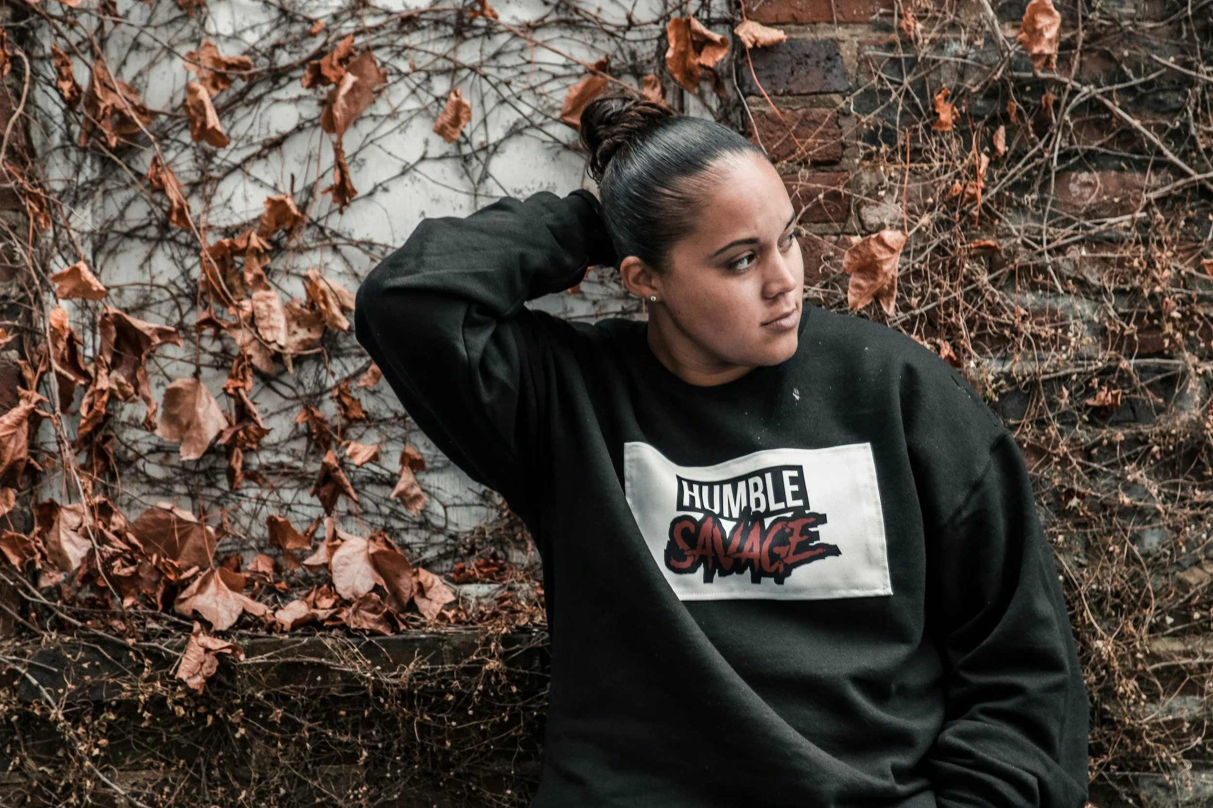 A woman with dark hair tied in a bun wearing a black sweatshirt with a graphic that says "Humble Savage" stands against a wall covered in dry, brown leaves and vines.