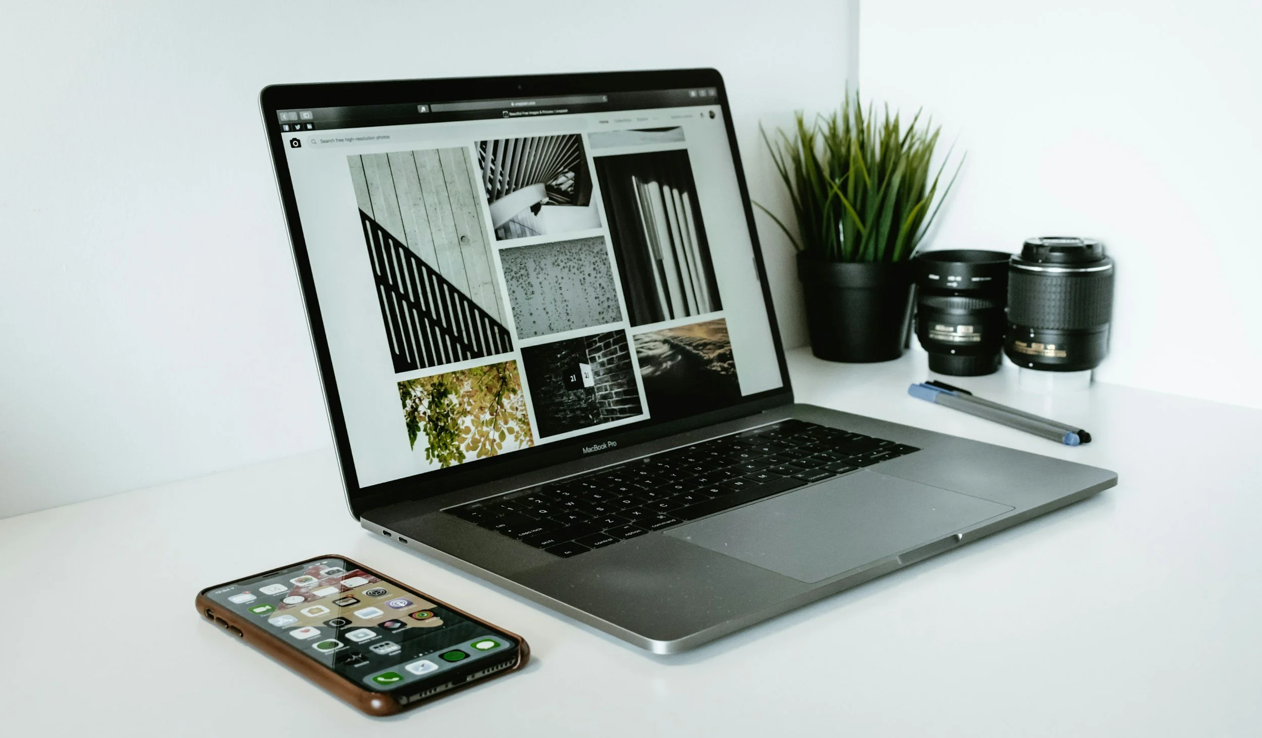 A MacBook Pro laptop displaying a collage of images on a white desk with a smartphone, a black plant pot with a green plant, and two camera lenses.