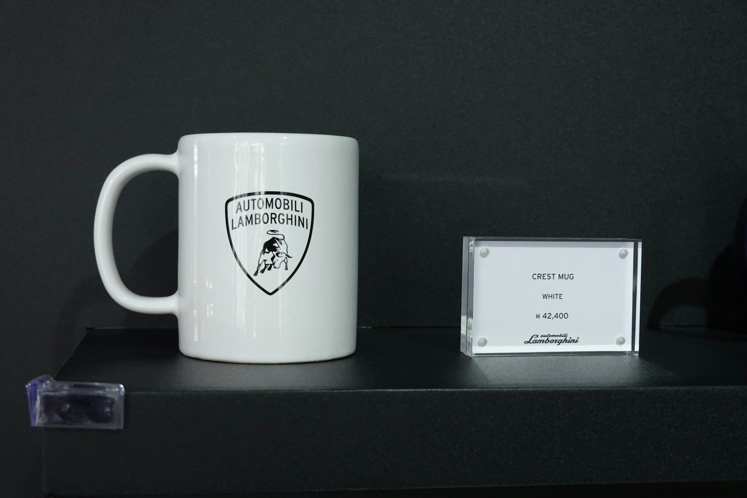 White ceramic mug with Lamborghini logo on display in a glass case at an exhibition, with a small placard indicating it is a 'Crest Mug' priced at 42,400 Korean won.