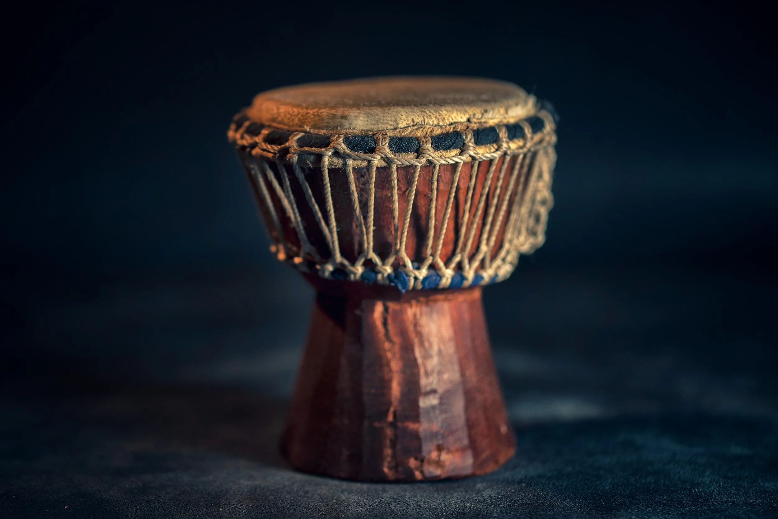 A traditional drum with a wooden body and a taut animal skin drumhead, surrounded by rope bindings, set against a dark background.